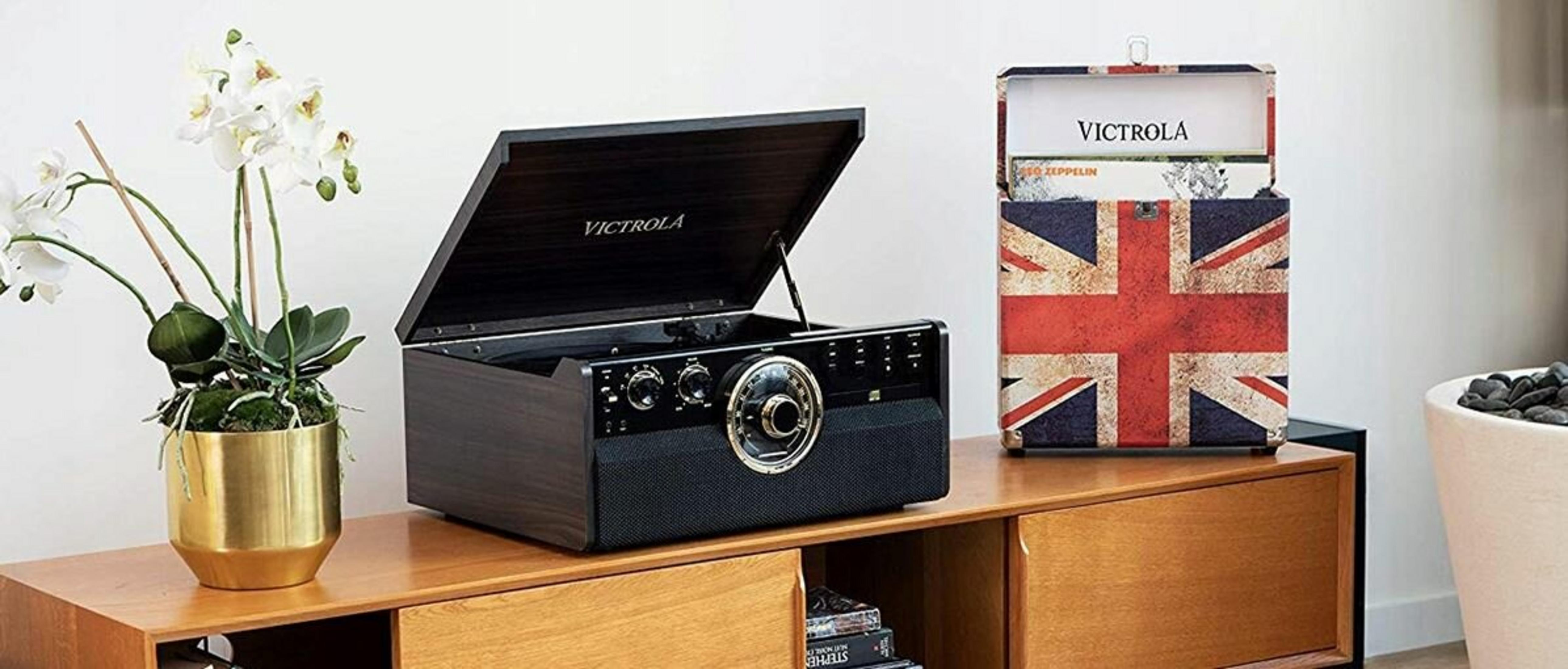 Un giradischi Victrola con il coperchio aperto e un portadischi Union Jack, con fiori bianchi in un vaso dorato.