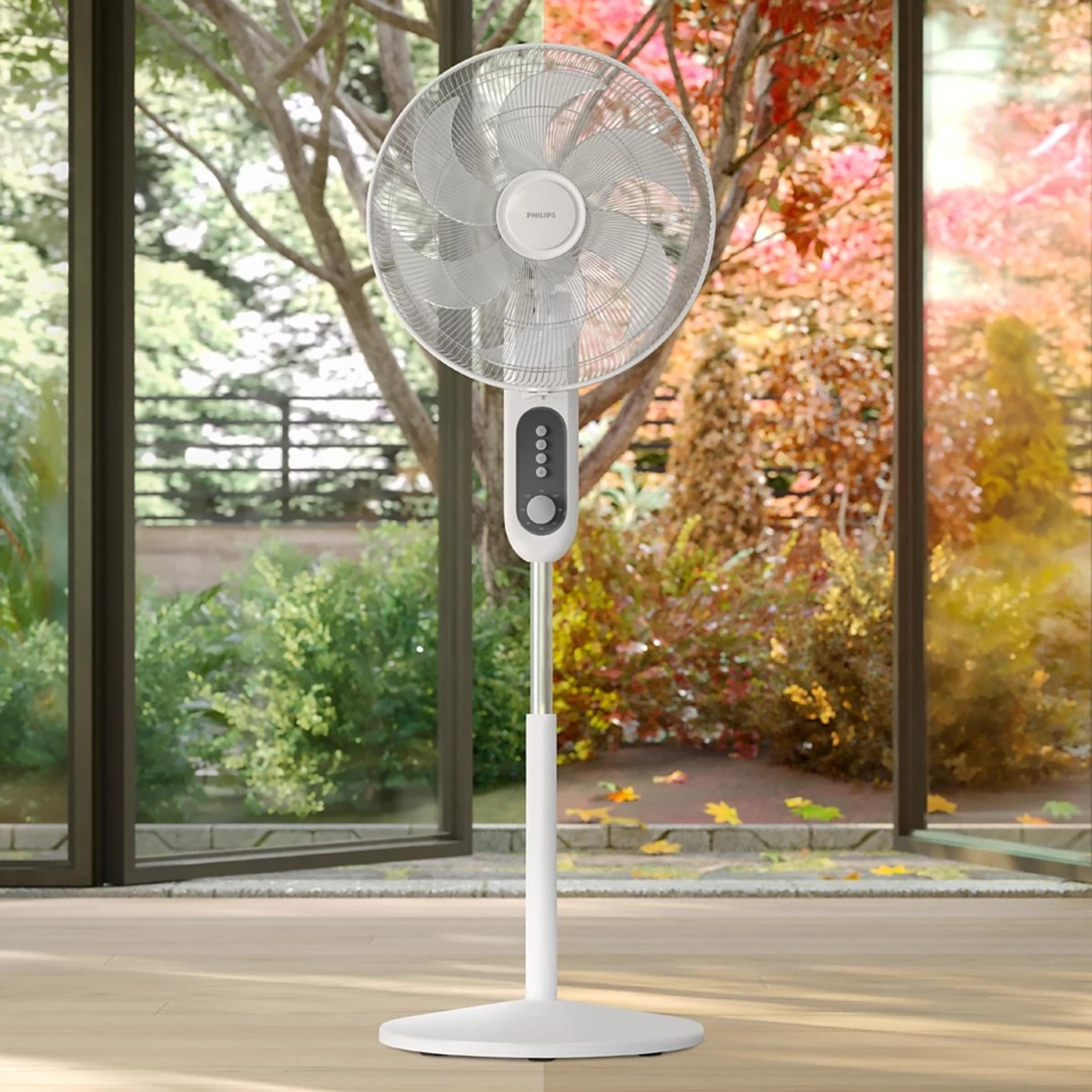 A white Philips fan stands on a wooden floor near a glass door. The background shows a garden with colorful trees.