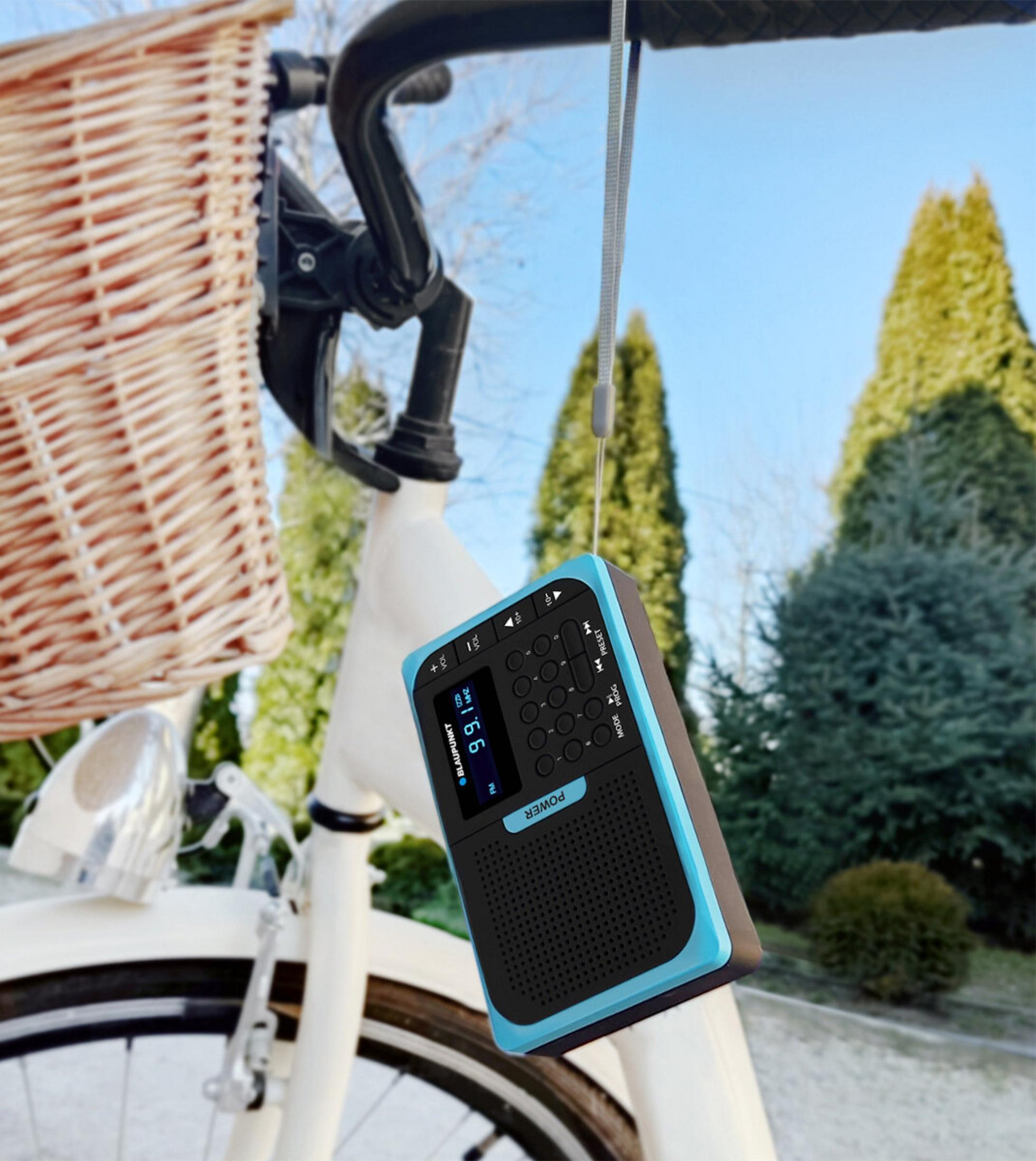 Una radio è appesa a una bici. È nera e blu. Un cestino è attaccato al manubrio della bici. Alberi e cielo sullo sfondo.