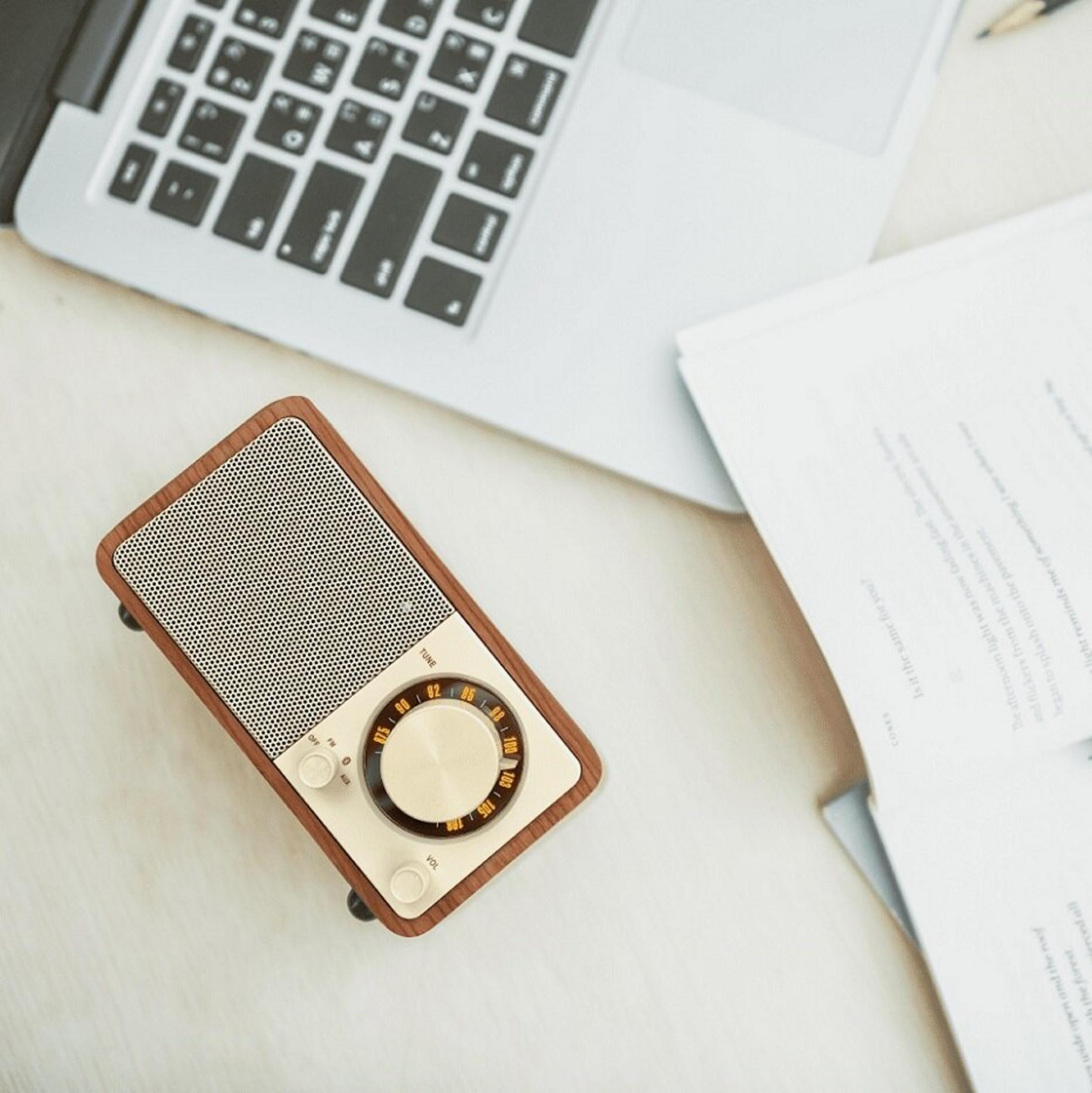 Zabytkowe radio, laptop i papiery na biurku. Radio ma drewnianą ramę, z pokrętłem. Laptop jest otwarty.