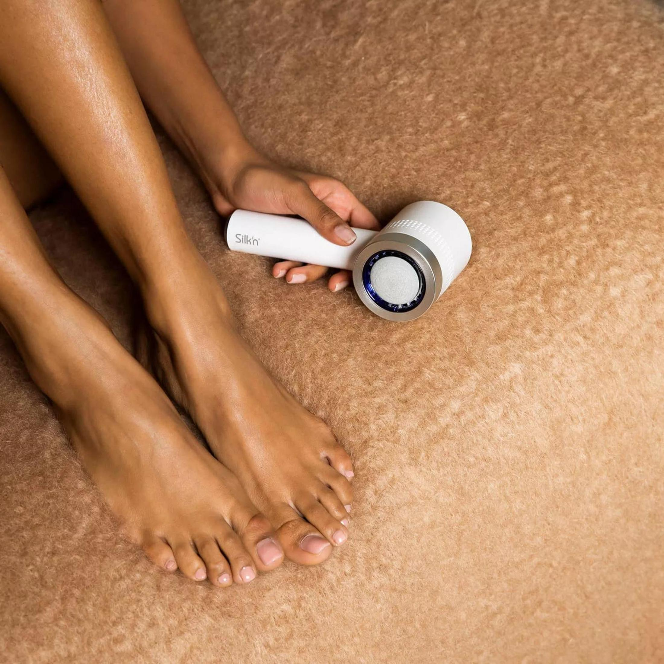 Osoba trzyma białe urządzenie Silkn w pobliżu stóp. Nogi i stopy widoczne są na brązowej, teksturowanej powierzchni.