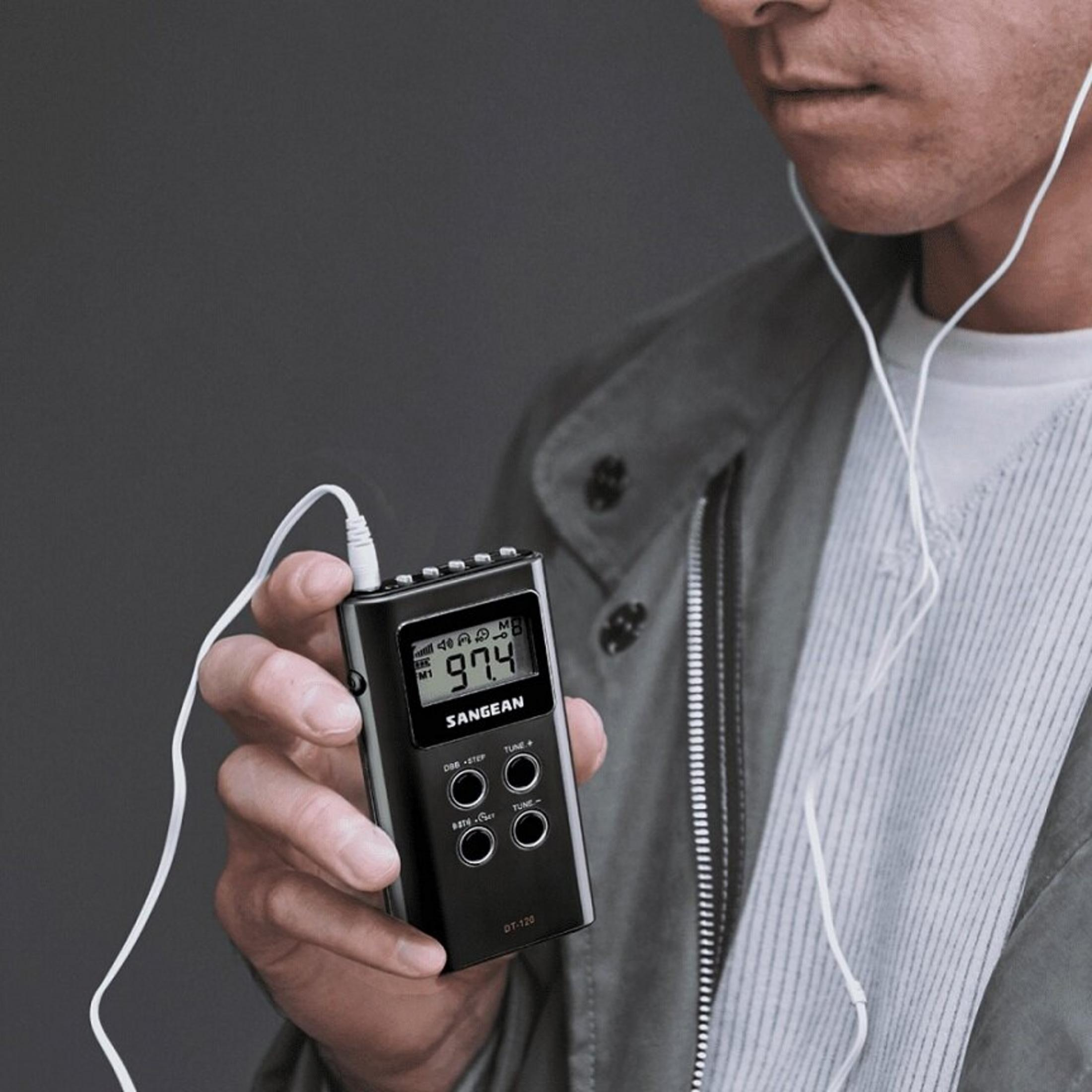 Un uomo tiene in mano una radio nera con display digitale che mostra 97.4. Auricolari bianchi collegati. Sfondo grigio.