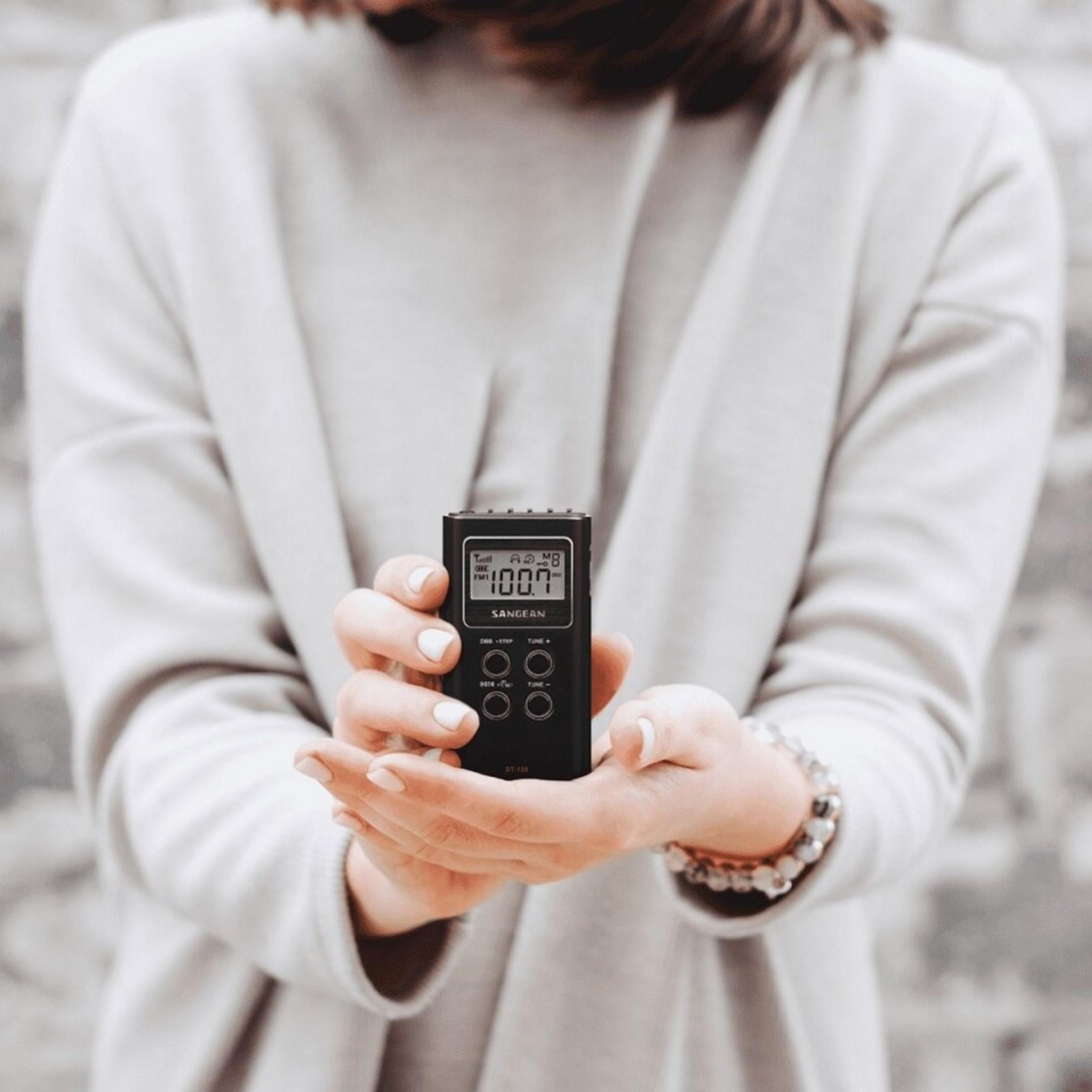 Una persona tiene in mano una radio nera Sangean BT-120. La radio mostra 100.7. La persona indossa un braccialetto e un maglione bianco.