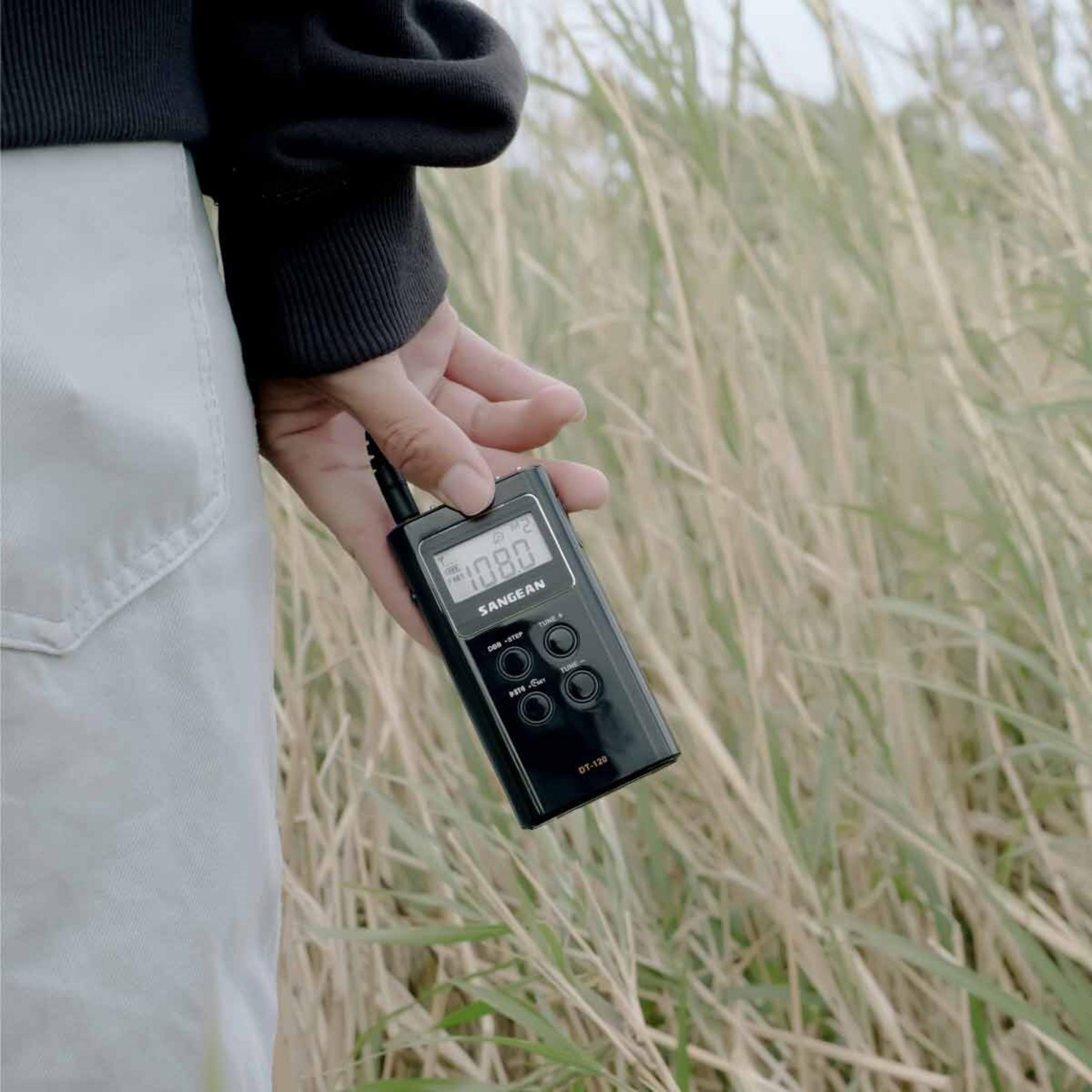Una mano tiene una radio nera Sangean DT-120 in un campo di erba alta e secca, persona che indossa un maglione nero e pantaloni chiari.
