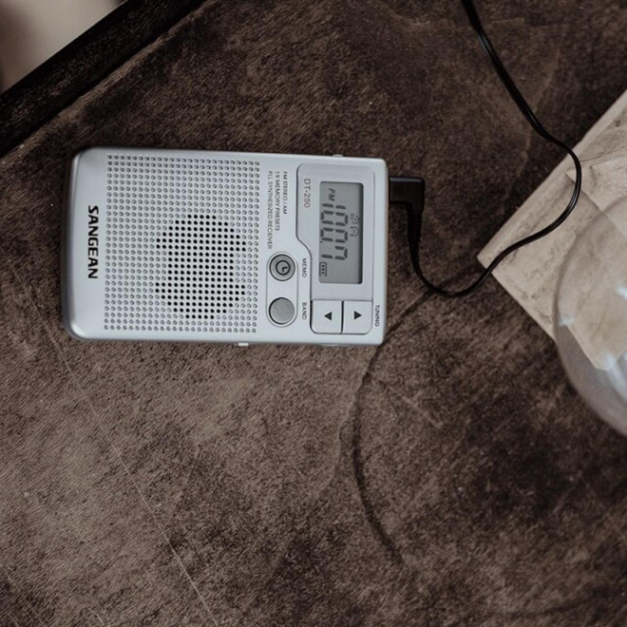 Srebrne radio SANGEAN DT-250 wyświetla 100.1 FM na ciemnej drewnianej powierzchni, z czarnym przewodem.