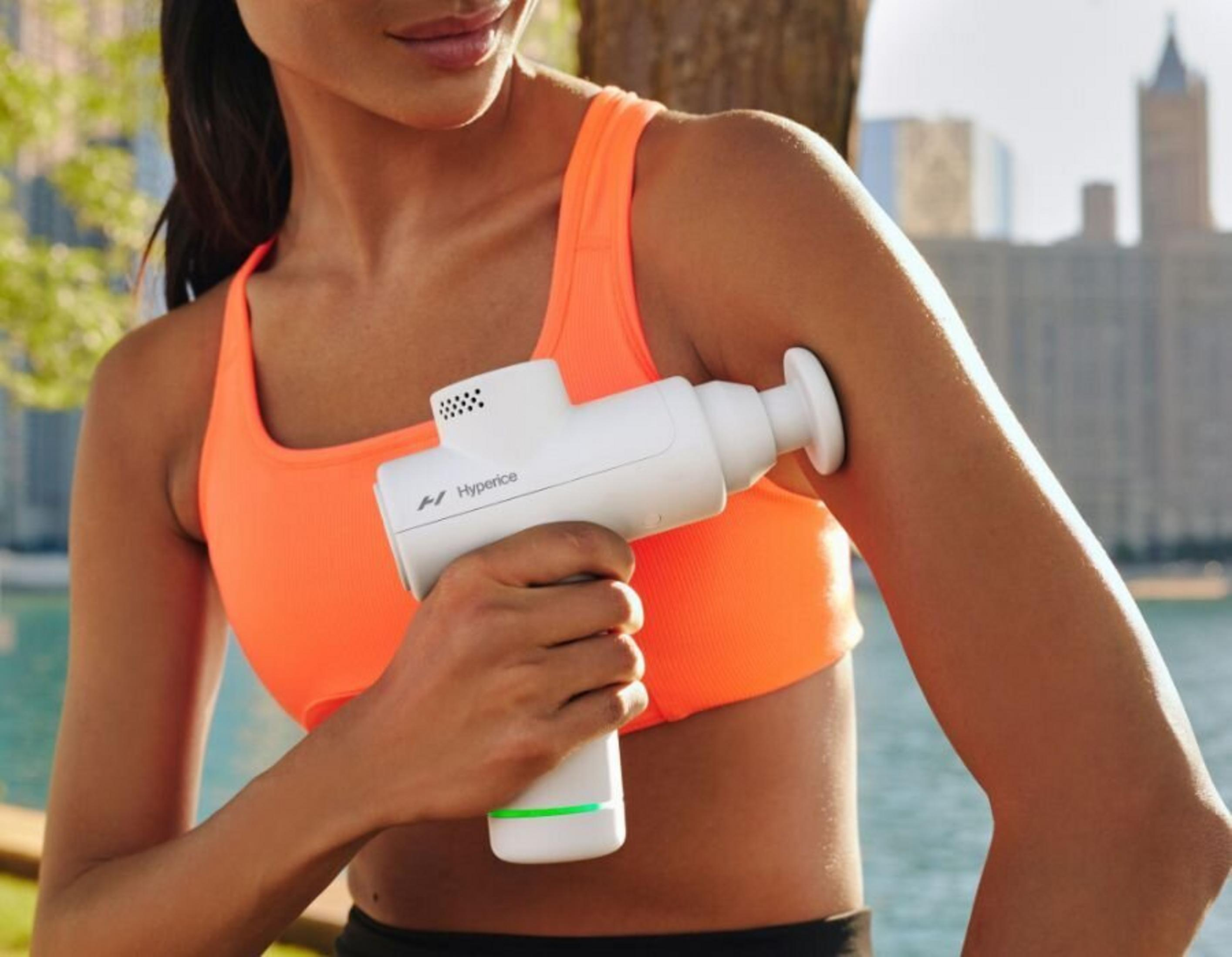 Una mujer con un sujetador deportivo naranja usa una pistola de masaje blanca en su brazo. Fondo: árboles y edificios.