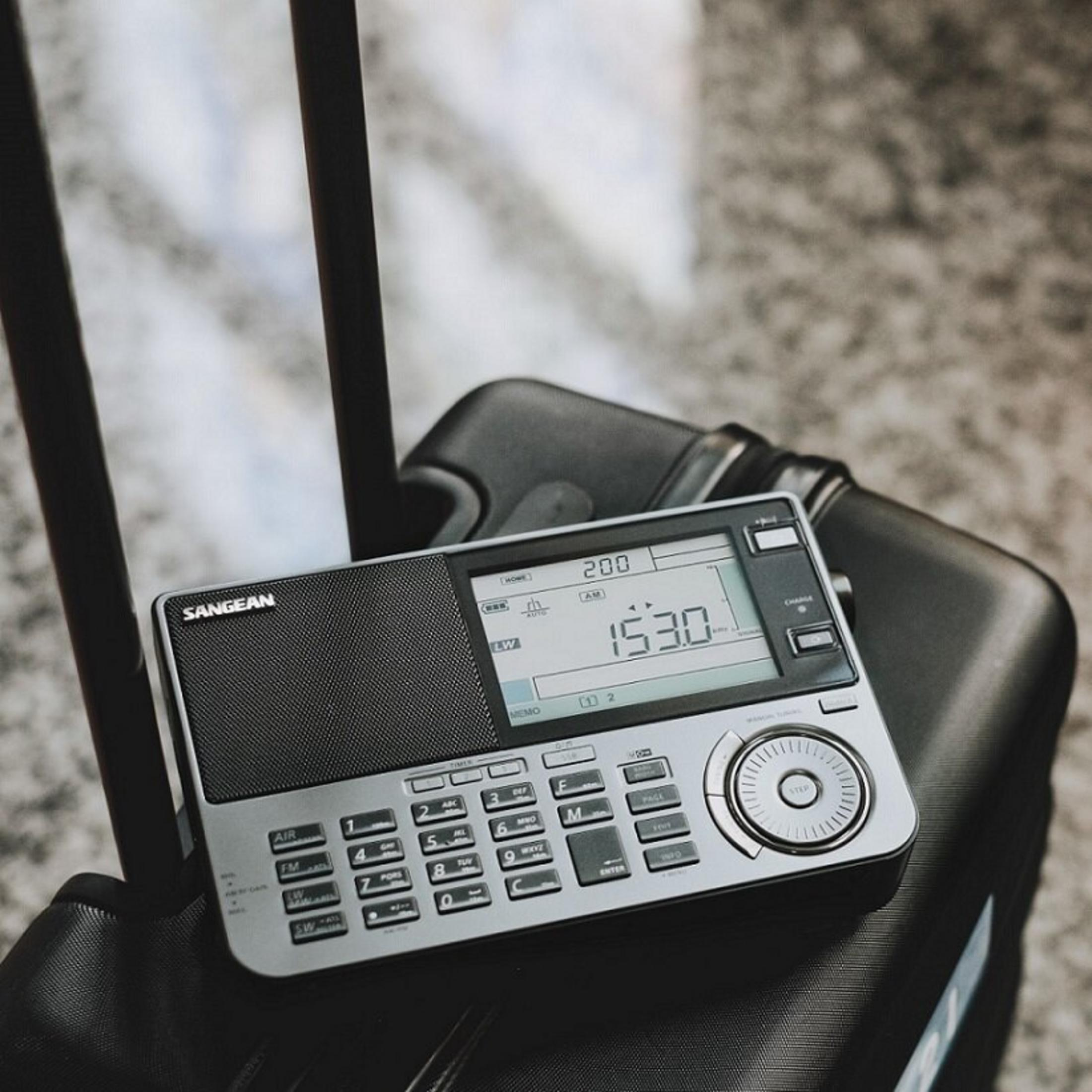 Srebrne radio SANGEAN wyświetla godzinę 15:30. Leży na czarnej walizce. Tło jest rozmyte.