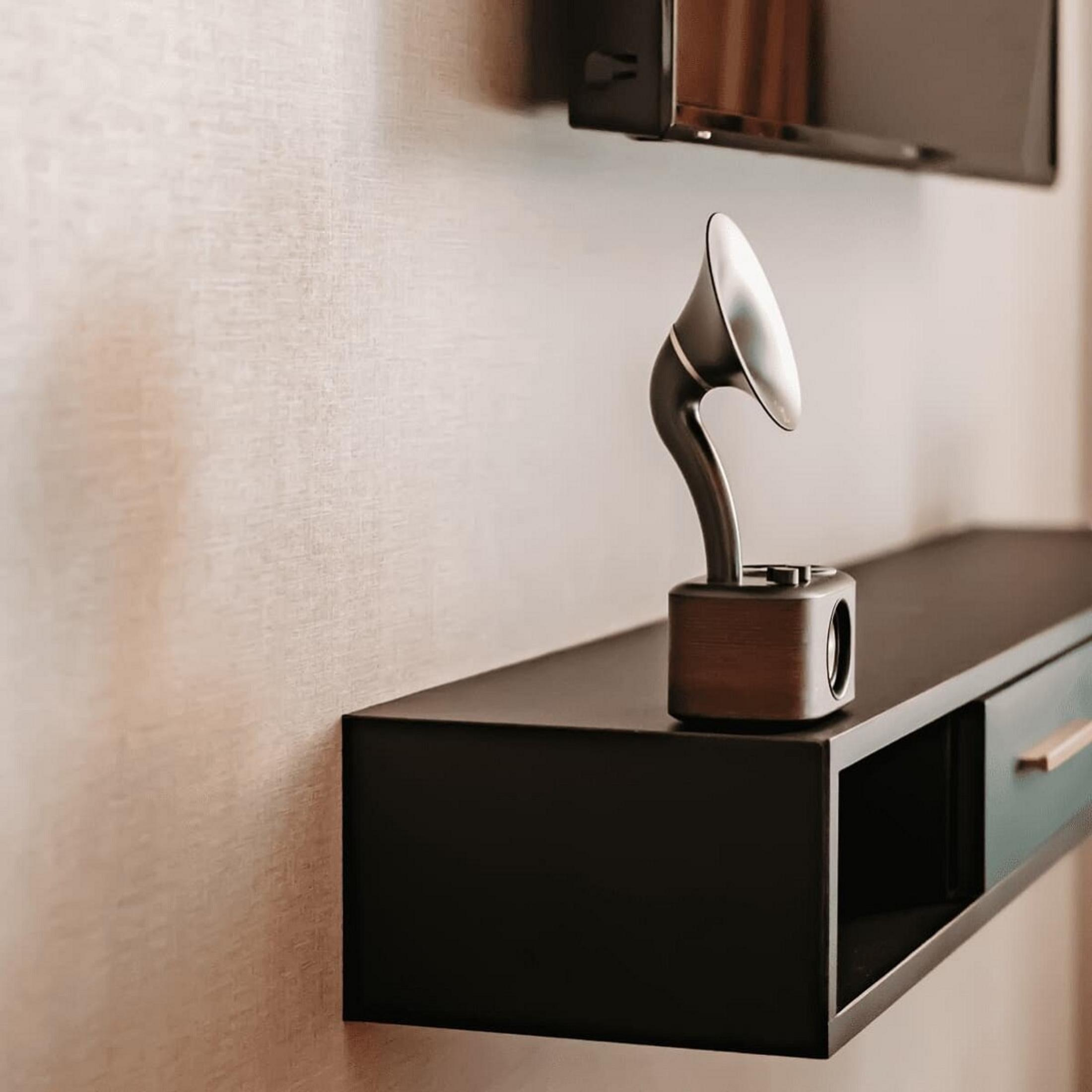 Un altavoz decorativo con forma de gramófono está sobre un estante negro. La pared es de color beige claro.