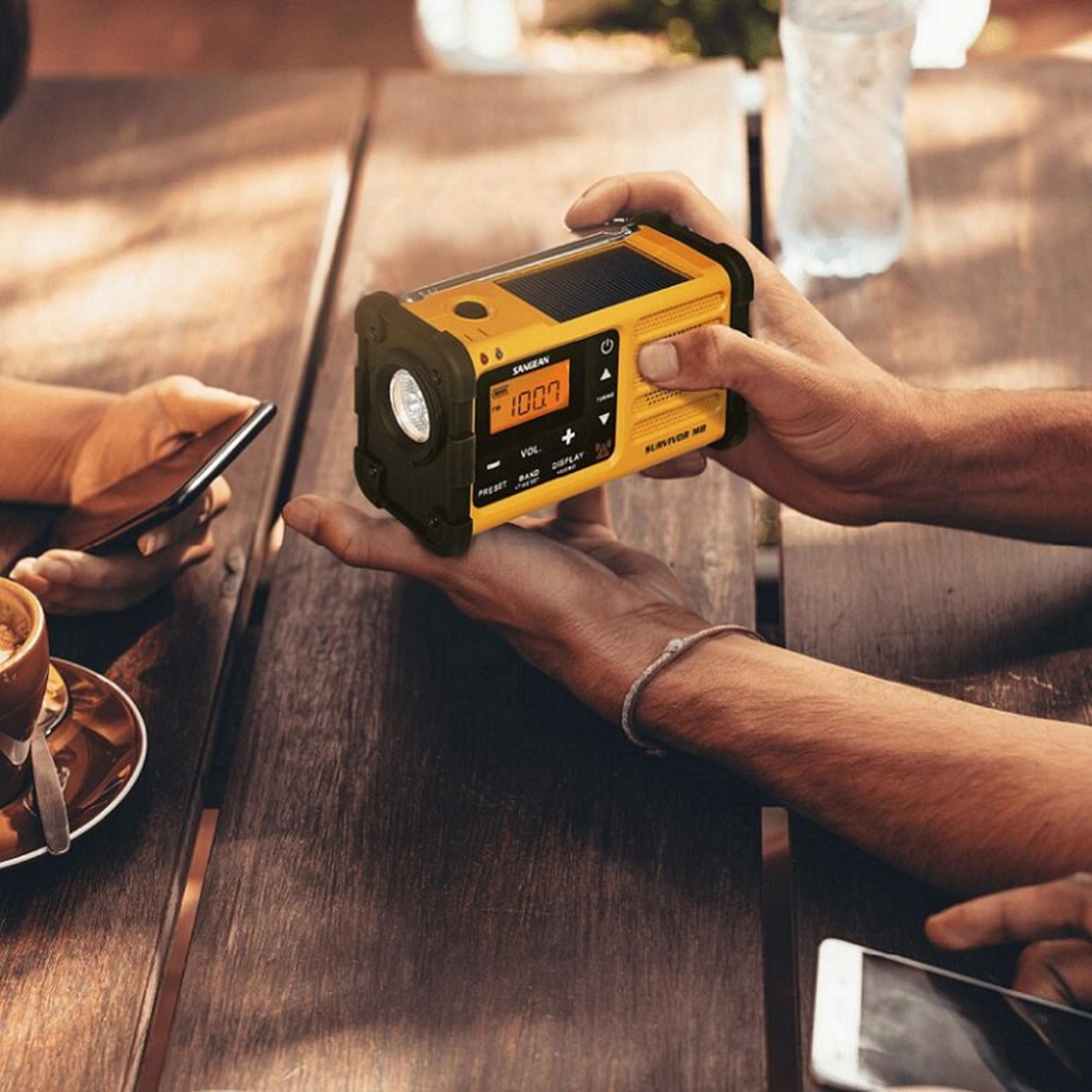 Una persona tiene in mano una radio gialla con una luce e un pannello solare. Un telefono, una tazza di caffè e una bottiglia d'acqua sono su un tavolo di legno.