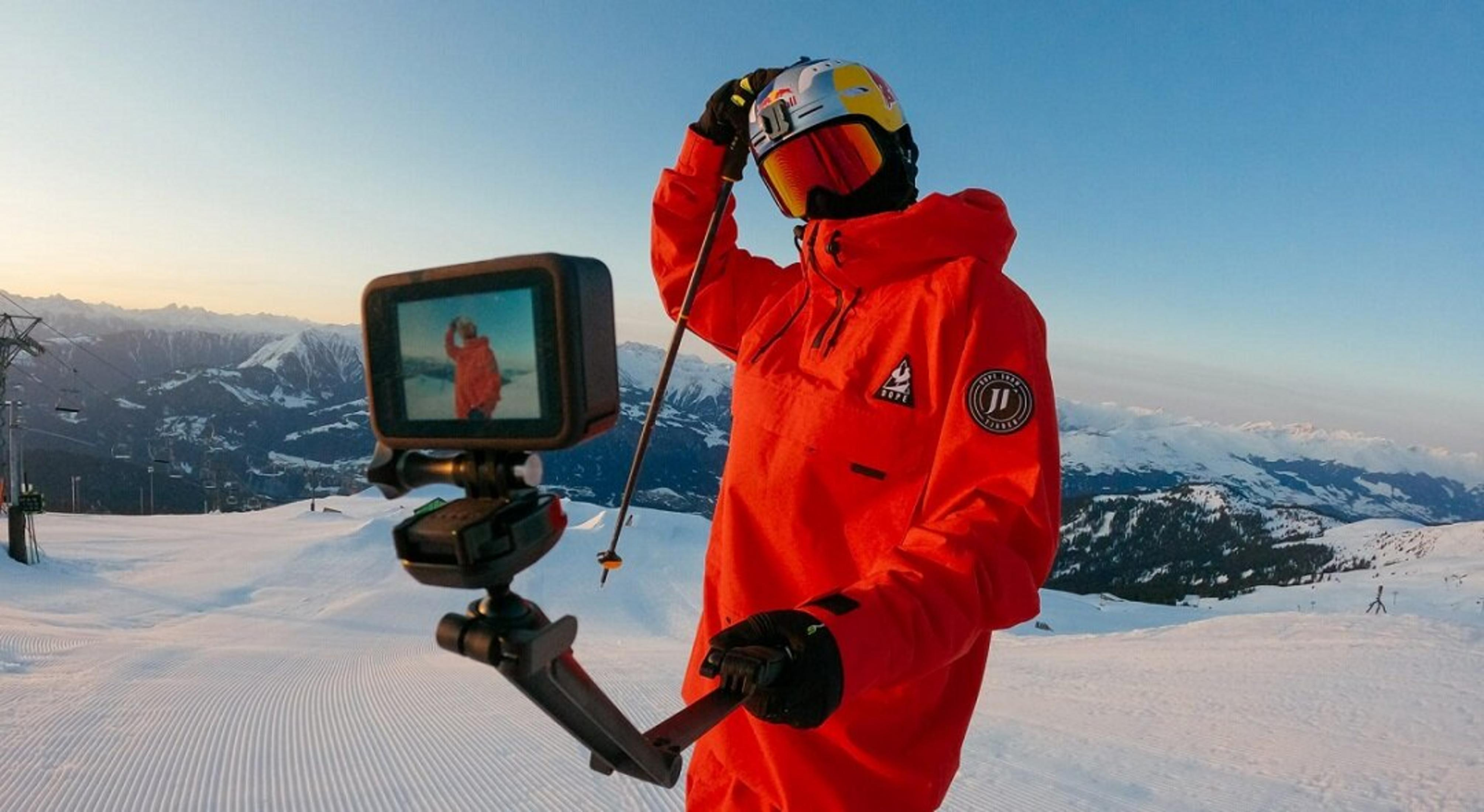 Un esquiador con chaqueta roja sostiene un palo selfie con una cámara, montañas al fondo, cielo azul.