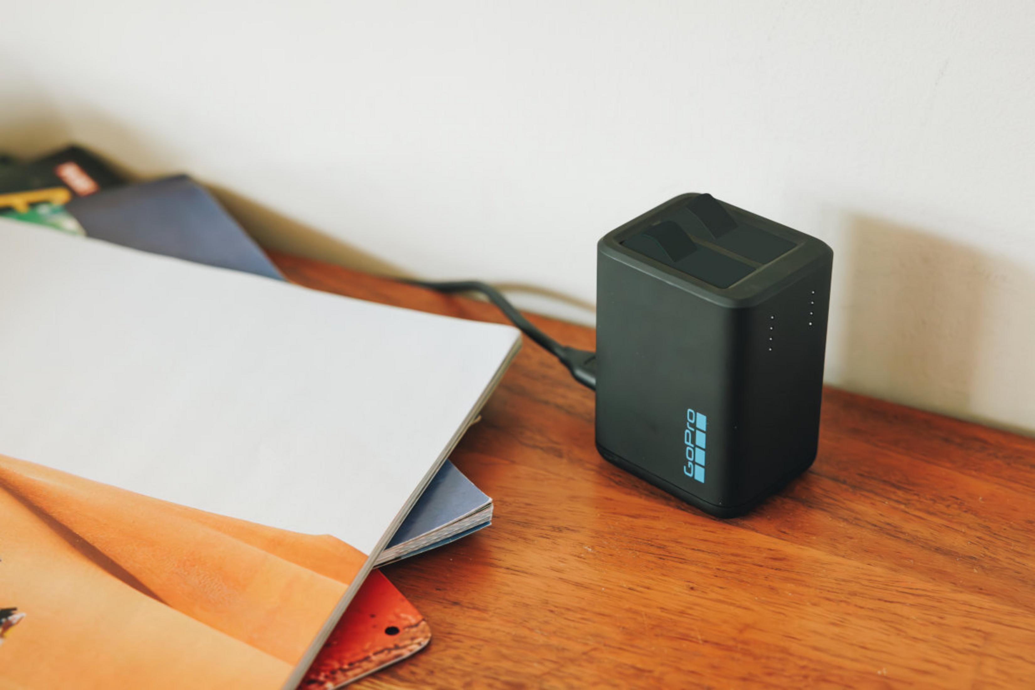 Czarna ładowarka GoPro stoi na drewnianej powierzchni obok książek. Biała ściana w tle.