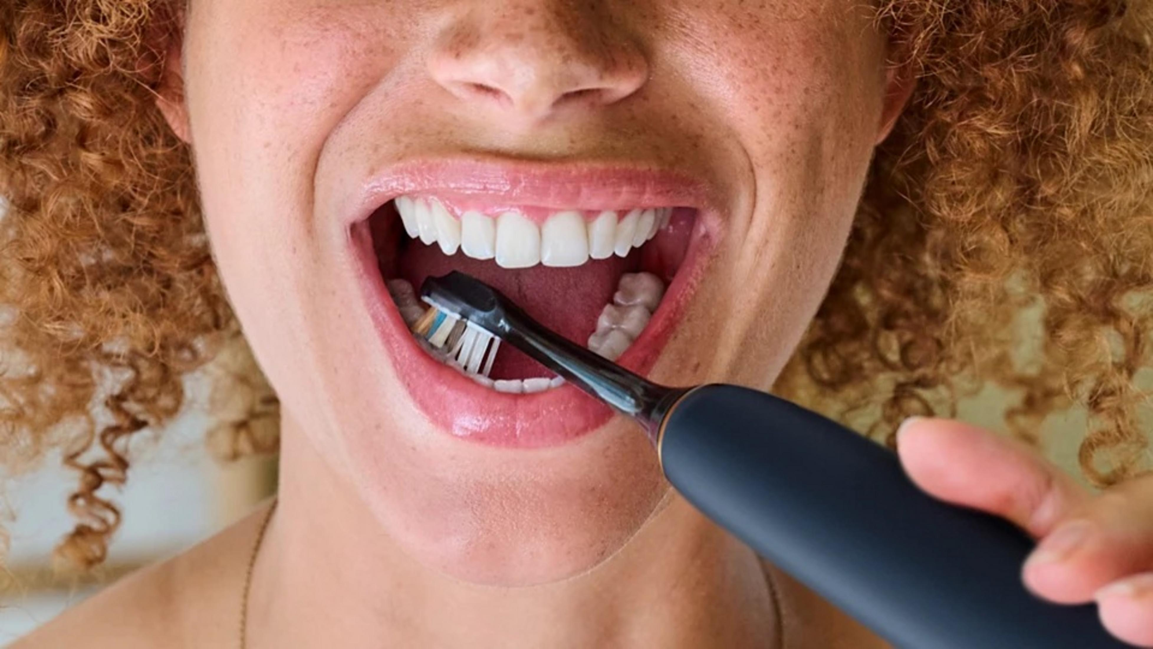 Una mujer se cepilla los dientes con un cepillo eléctrico. Tiene la boca abierta, revelando dientes blancos. El pelo rizado enmarca su rostro.
