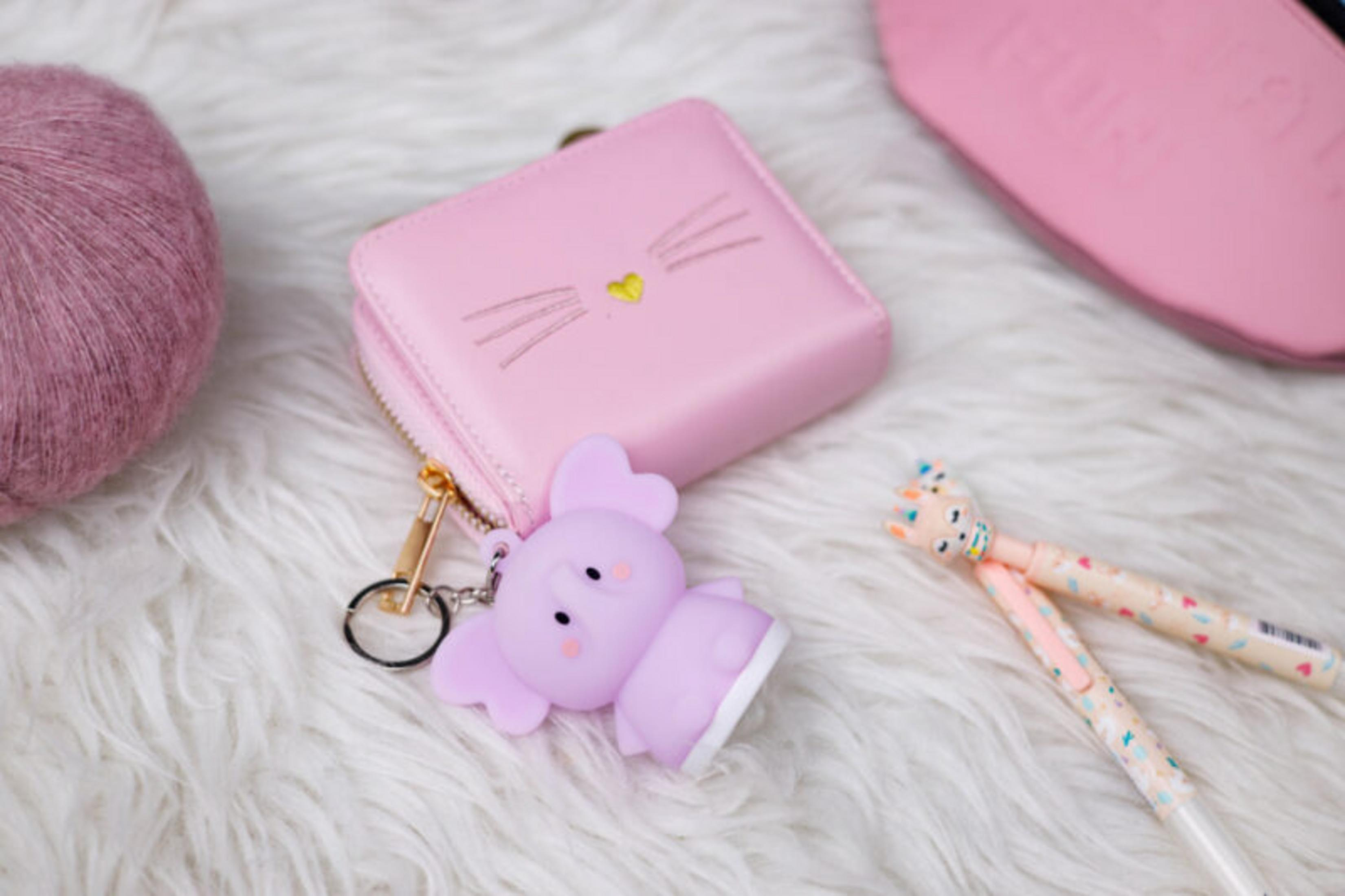 Artículos rosados sobre piel blanca: cartera, llavero de elefante, bolígrafos y ovillo de lana. La cartera tiene un diseño de cara de gato.