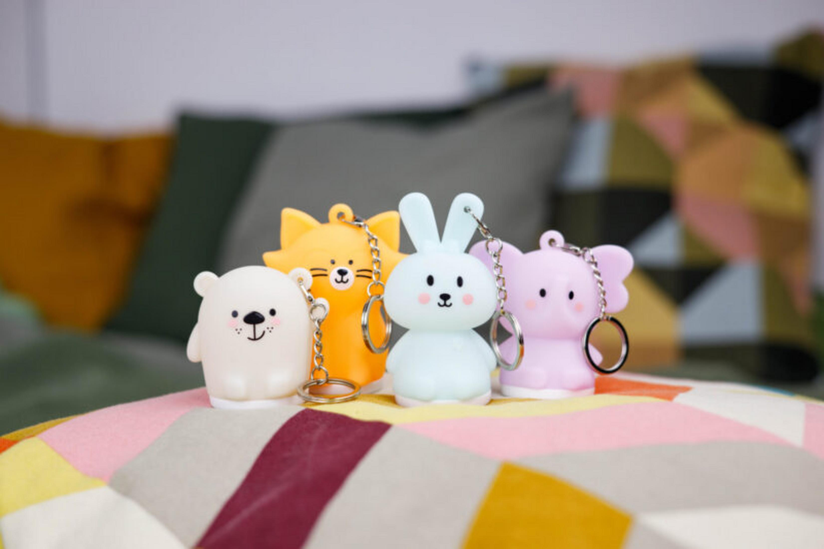 Cuatro llaveros de animales: oso, gato, conejo y elefante. Están sobre una superficie estampada, con un fondo borroso de almohadas.