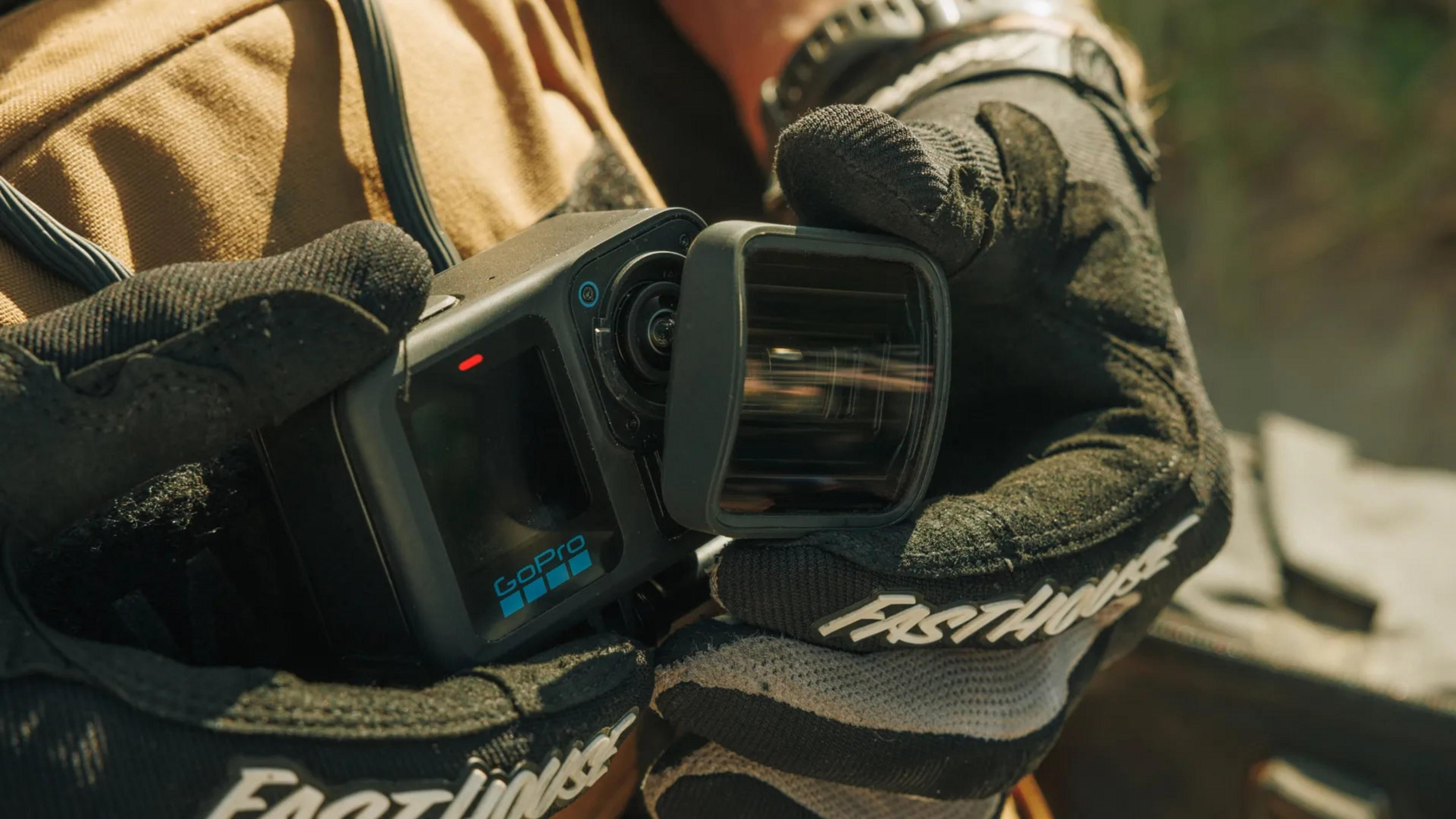 Eine Person mit schwarzen Handschuhen hält eine GoPro-Kamera. Die Kamera ist schwarz mit blauem GoPro-Logo. Der Hintergrund ist unscharf.