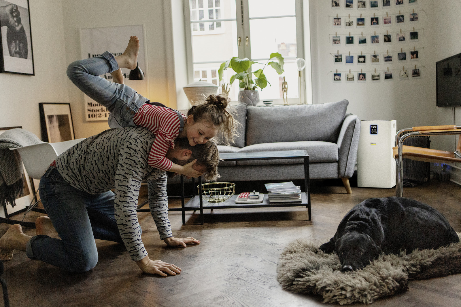 Ein Mann kniet mit einem Mädchen auf dem Rücken. Ein schwarzer Hund liegt auf einem Teppich. Eine Couch, ein Fenster und ein Luftreiniger sind im Hintergrund.