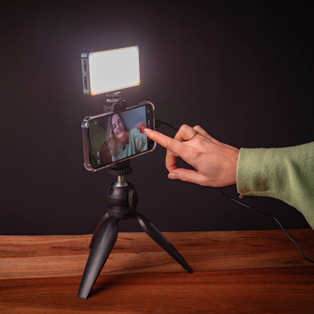 A woman's hand touches a phone screen displaying a person. The phone is on a tripod with a light. Background is a black wall and wood table.