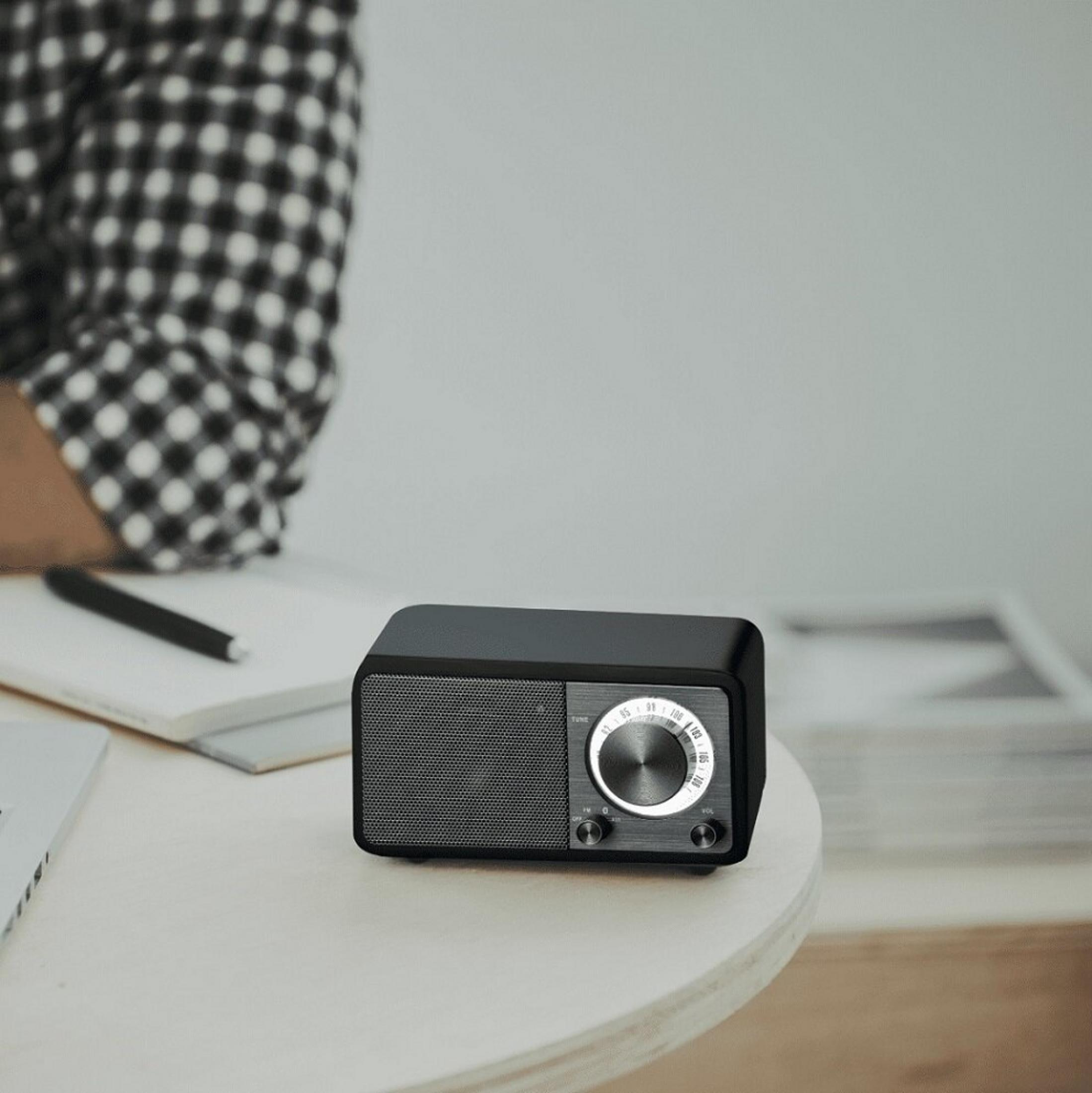 Czarny radioodbiornik na białym stole. Częściowo widoczna osoba w koszuli w kratę w tle.
