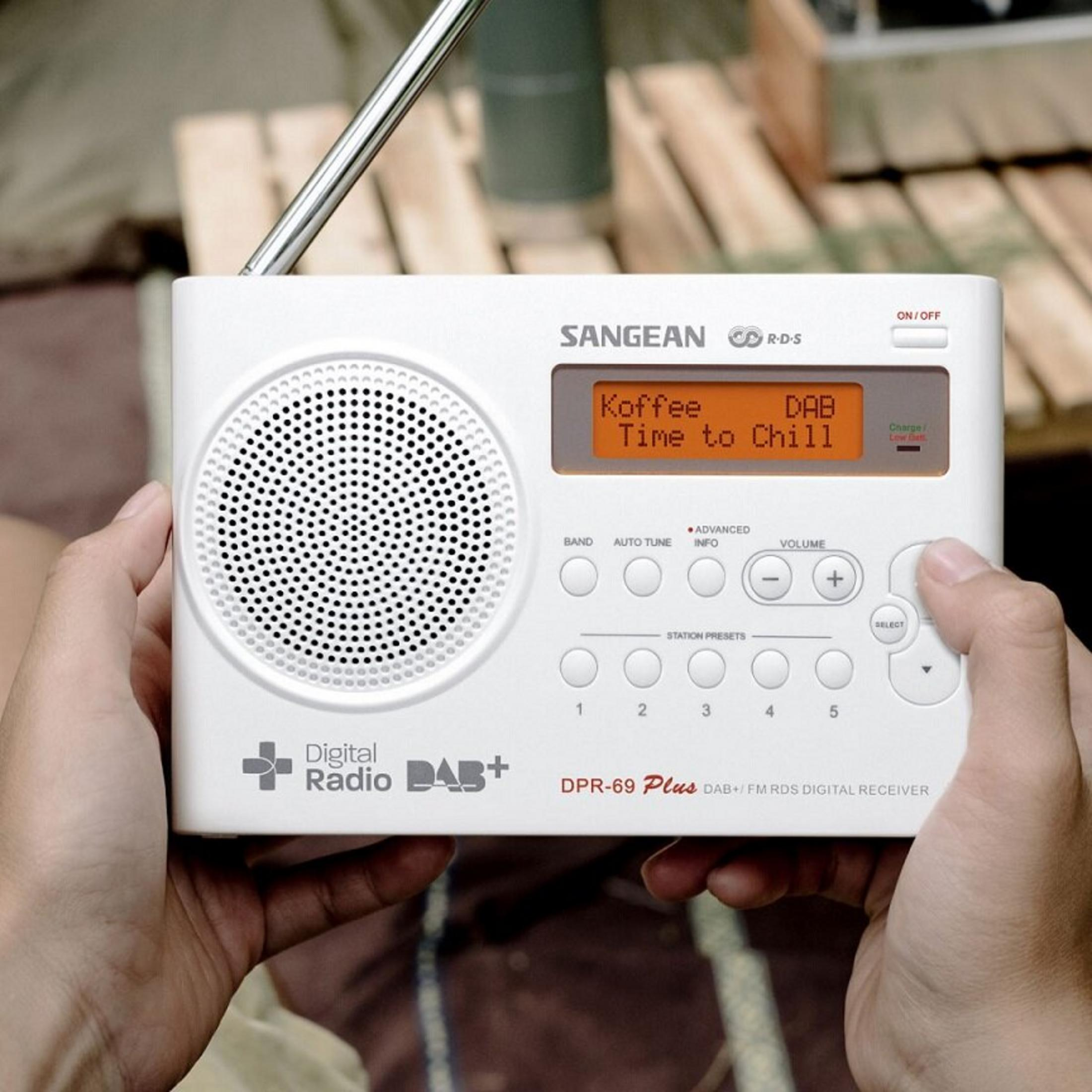 Ein weißes Digitalradio wird von Händen gehalten. Der Bildschirm zeigt 'Koffee Time to Chill'. Das Radio hat Knöpfe und einen Lautsprecher.