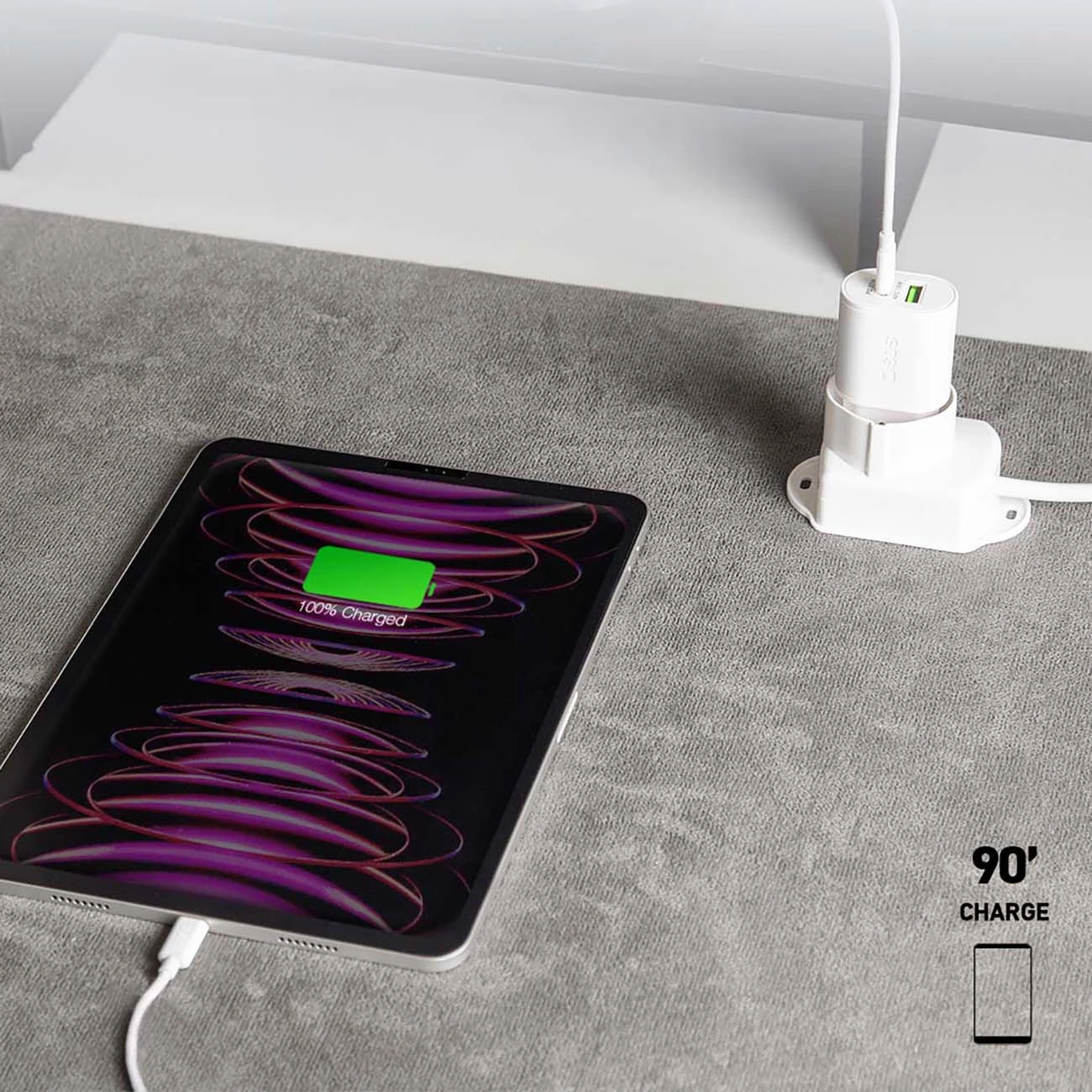 Un iPad si sta caricando, mostrando 100% di carica. È collegato a un caricabatterie bianco su una superficie grigia. Lo sfondo è bianco.