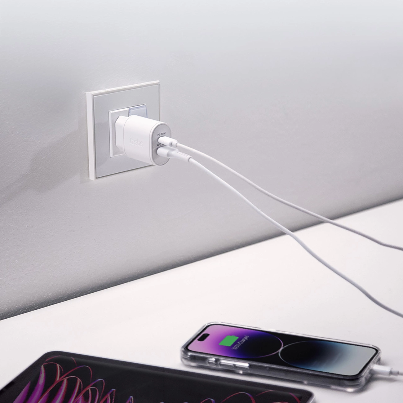 Caricabatterie bianco collegato a una presa a muro, che carica un telefono su una superficie bianca. È presente anche un altro dispositivo.