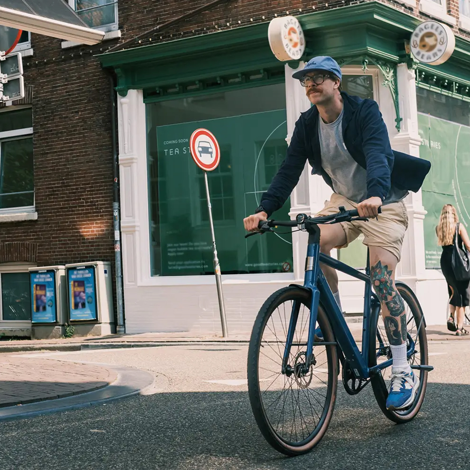 Ein Mann auf einem blauen Fahrrad fährt eine Straße entlang. Er trägt eine Kappe und hat Tätowierungen auf dem Bein. Gebäude und Straßenschilder sind im Hintergrund.
