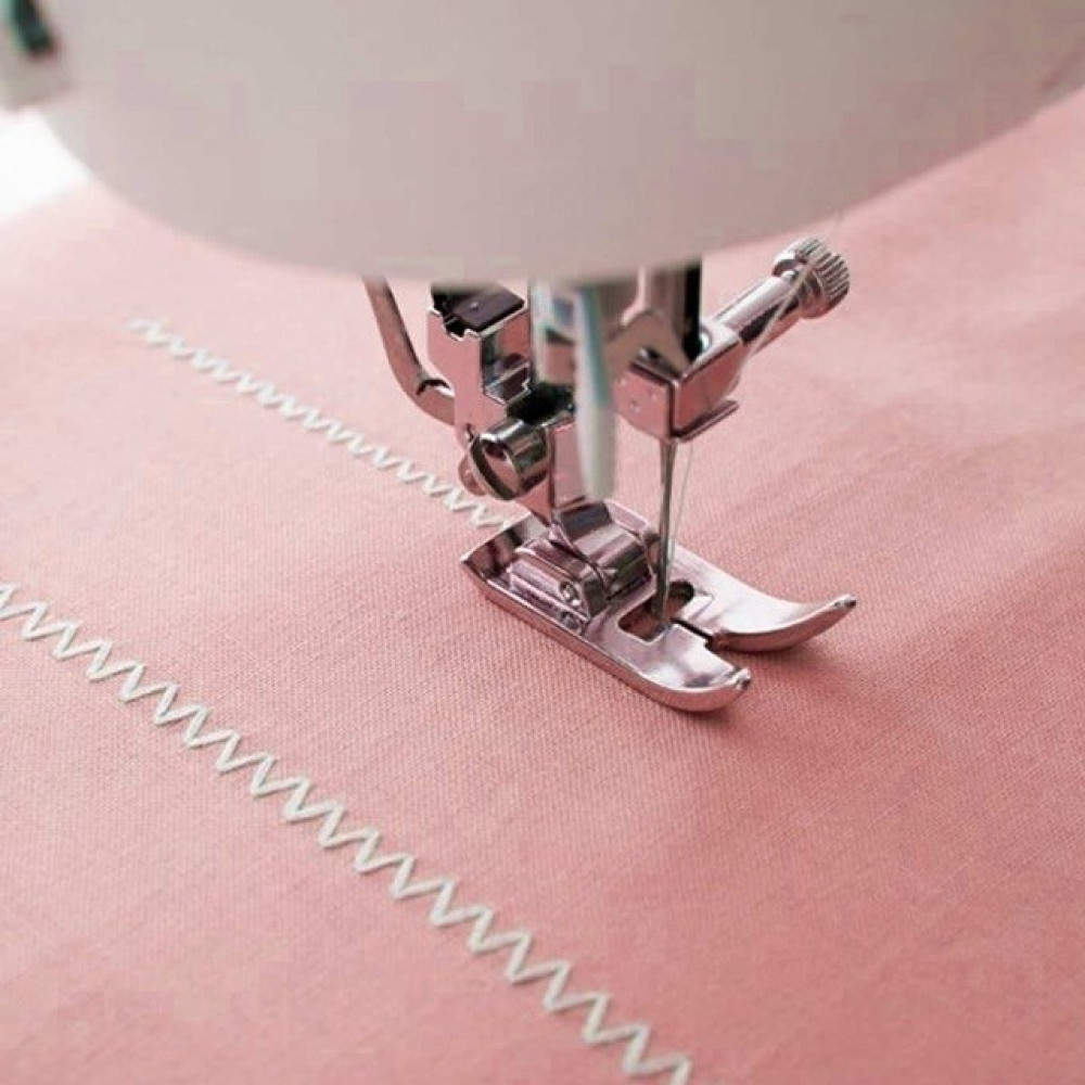 Igła maszyny do szycia zszywa białą nić na różowej tkaninie. Widoczna jest stopka maszyny oraz wzór materiału.