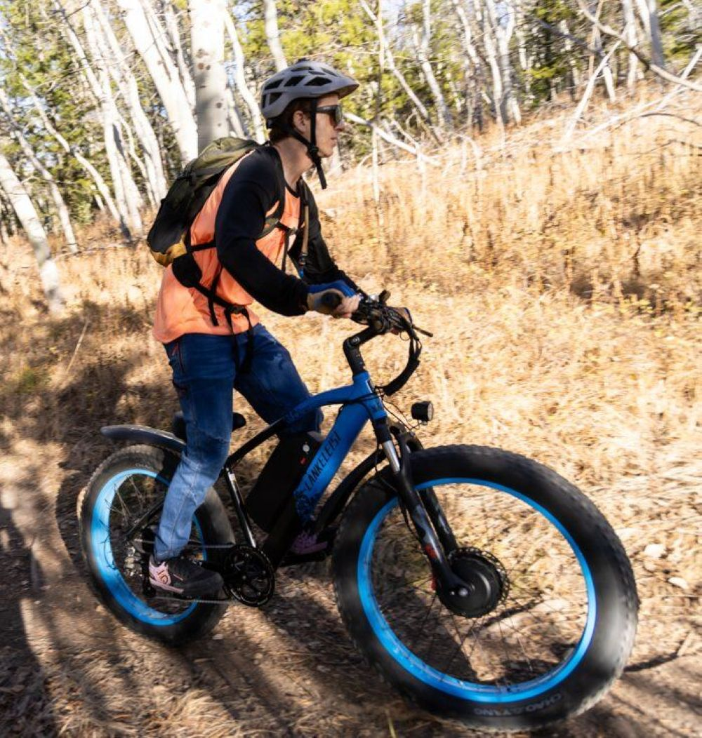 Una persona monta una bicicleta eléctrica azul en un camino de tierra en un bosque. Lleva casco, gafas de sol y chaleco naranja.