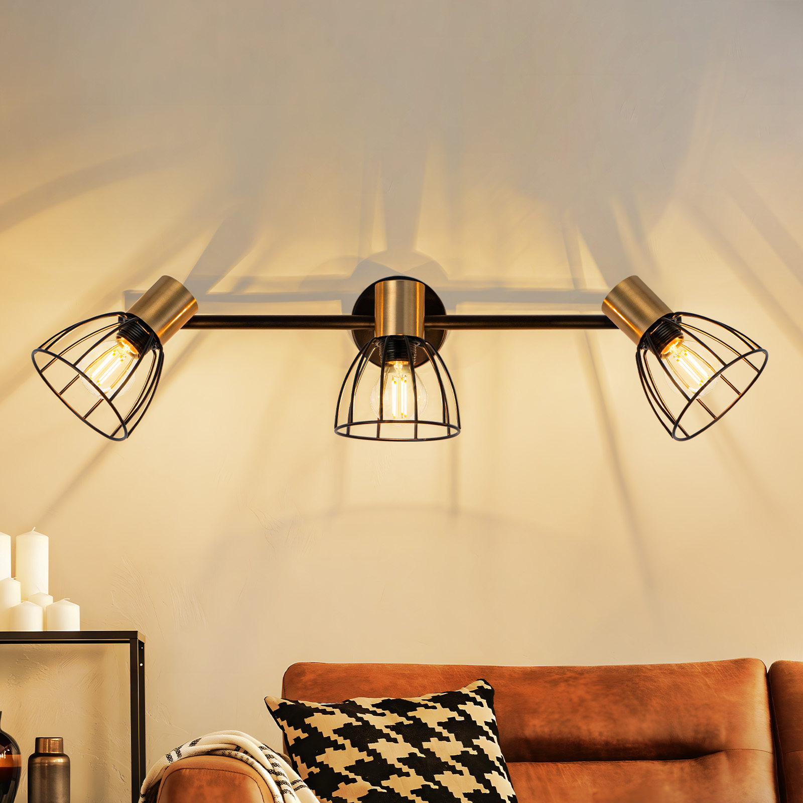 Una lampada moderna con tre luci. Sotto, un divano in pelle con un cuscino fantasia.