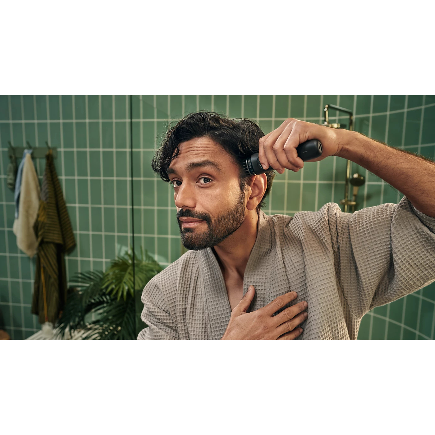 Mężczyzna w szlafroku goli ucho czarną trymerką w łazience z zielonymi kafelkami.