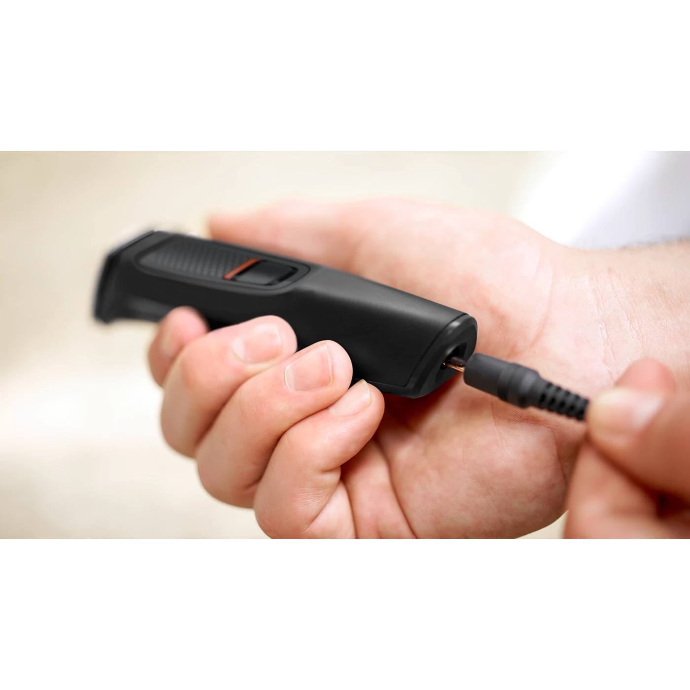 A person is plugging a black trimmer into a charging cable. The trimmer has an orange button.