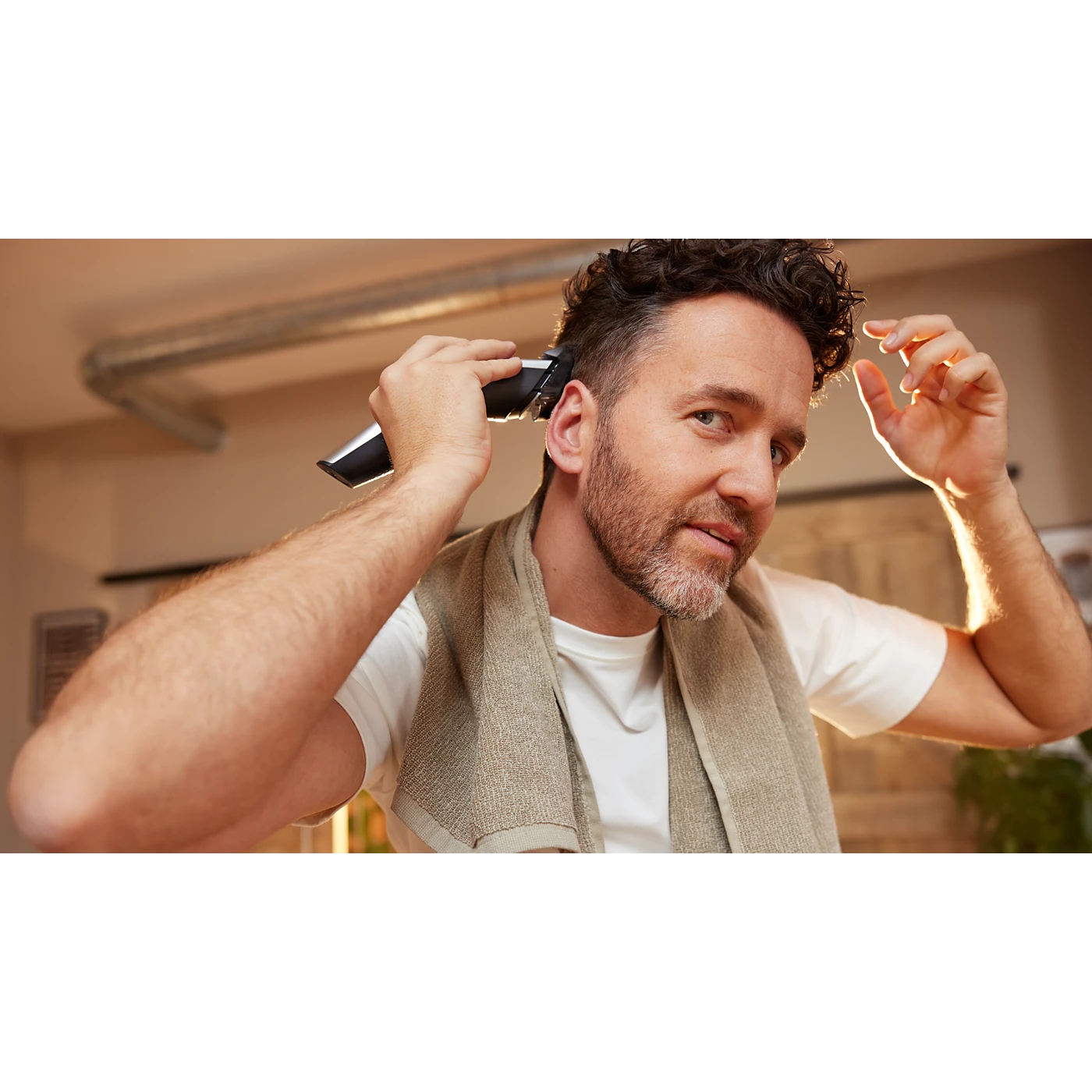 Hombre corta su cabello con recortadora. Viste camisa blanca y tiene toalla al cuello. Fondo borroso.