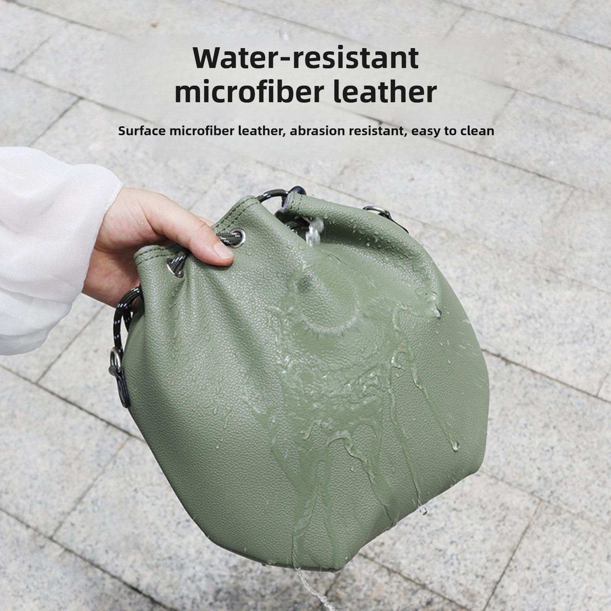 Eine grüne Ledertasche wird von einer Hand gehalten. Wasser wird auf die Tasche gegossen, Wassertropfen auf Stein.
