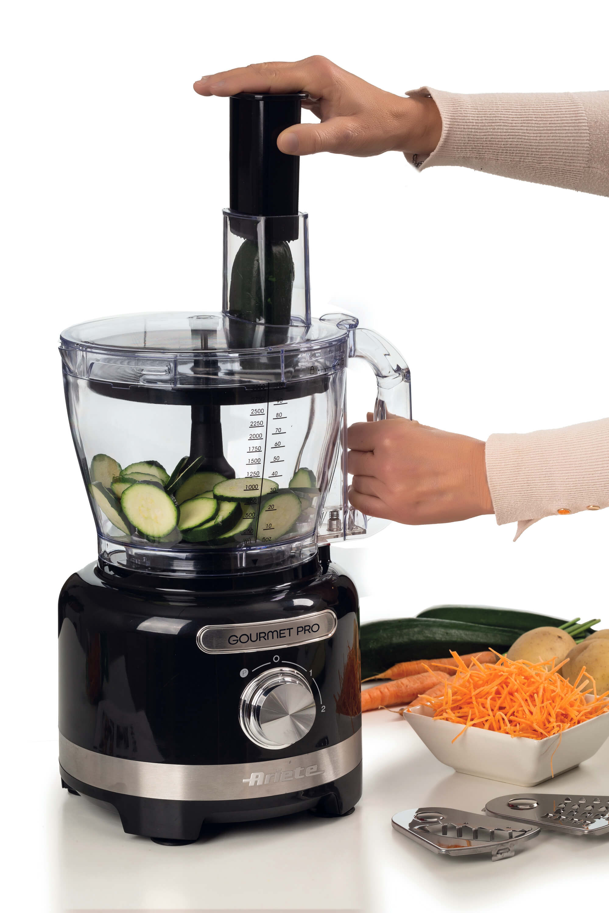 Un robot da cucina con base nera e contenitore trasparente. Una mano mette zucchine sopra.