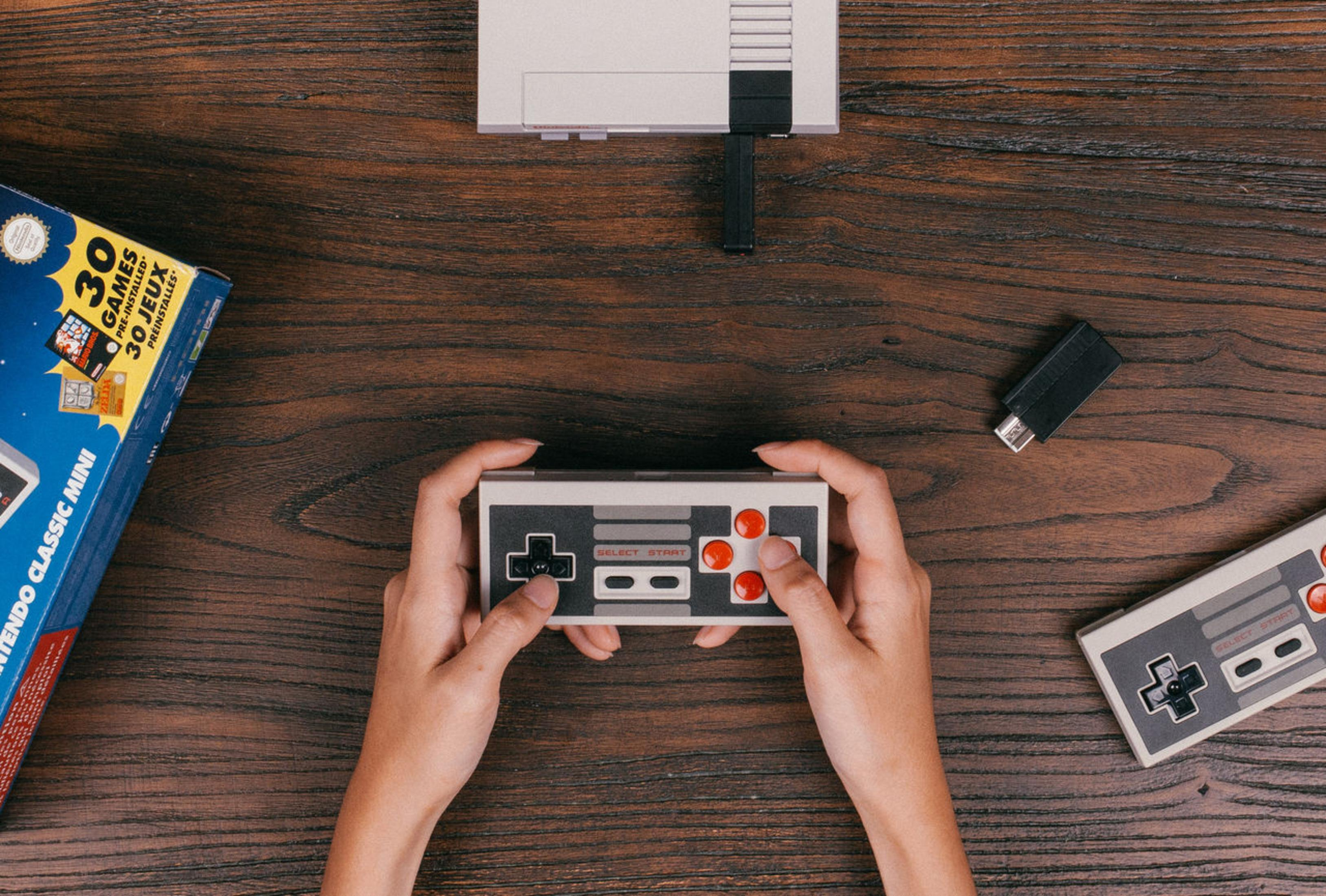 Mani che tengono un controller NES su un tavolo di legno, con console, scatola di gioco e controller extra.