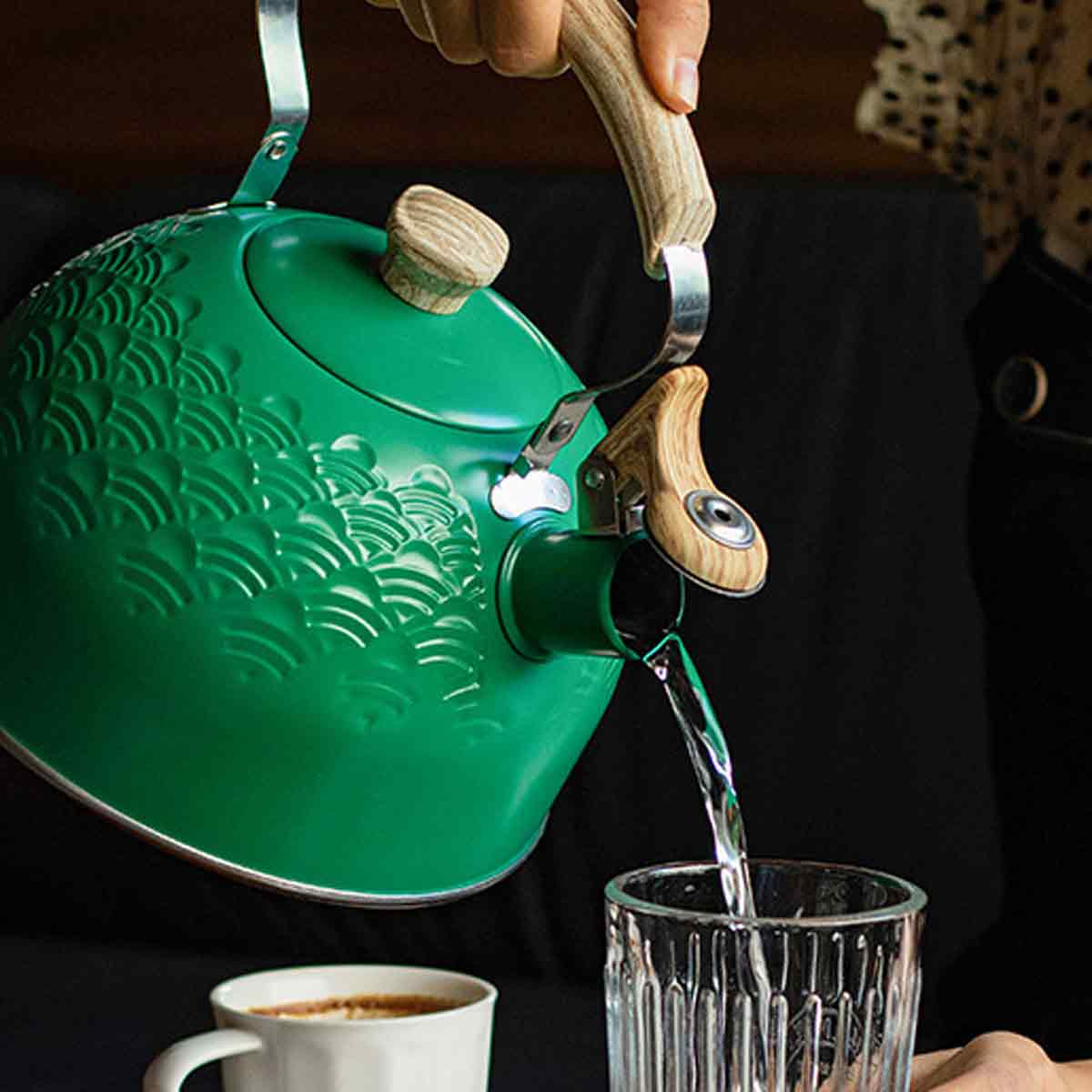 Ein grüner Wasserkocher gießt Wasser in ein Glas. Eine weiße Tasse mit Kaffee befindet sich im Vordergrund. Der Wasserkocher hat einen Holzgriff.