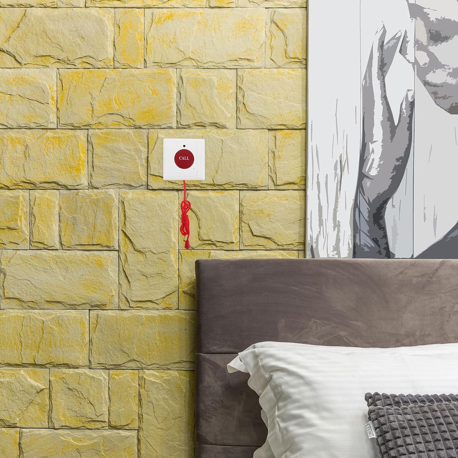 Eine Schlafzimmerwand mit gelben Steinziegeln, einem Rufknopf und einem Gemälde. Ein Bett mit weißem Kissen und grauem Kopfteil im Vordergrund.
