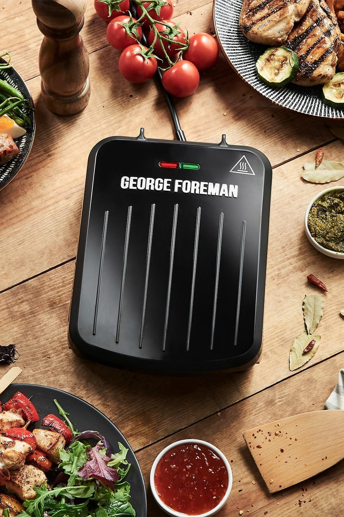 Ahşap bir masada George Foreman ızgarası, yemek ve domatesler. Yemek, ızgara tavuk, sebzeler ve sosları içerir.