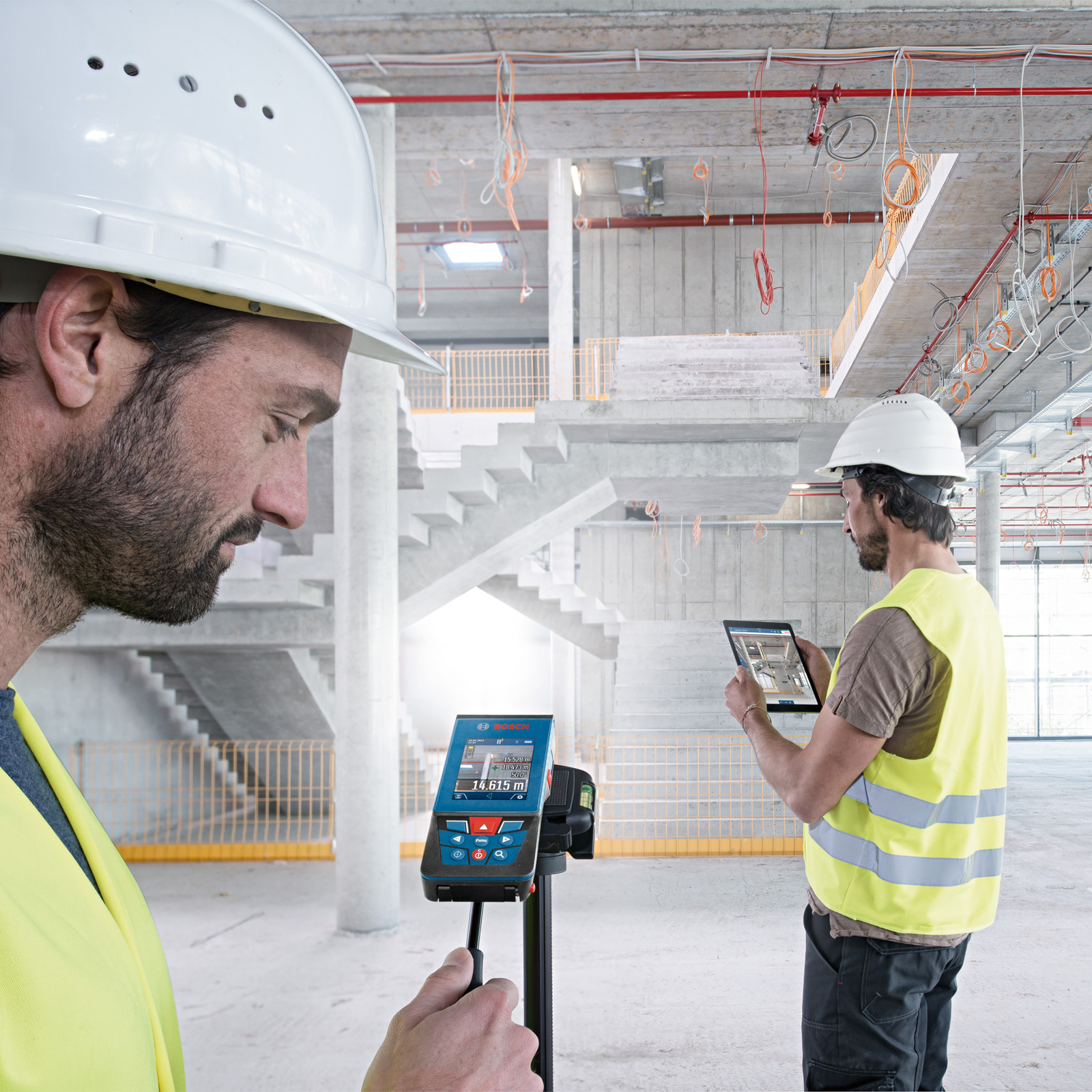 Bir inşaat alanında iki inşaat işçisi. Biri ölçüm cihazı, diğeri tablet kullanıyor. Beton yapı görünür durumda.