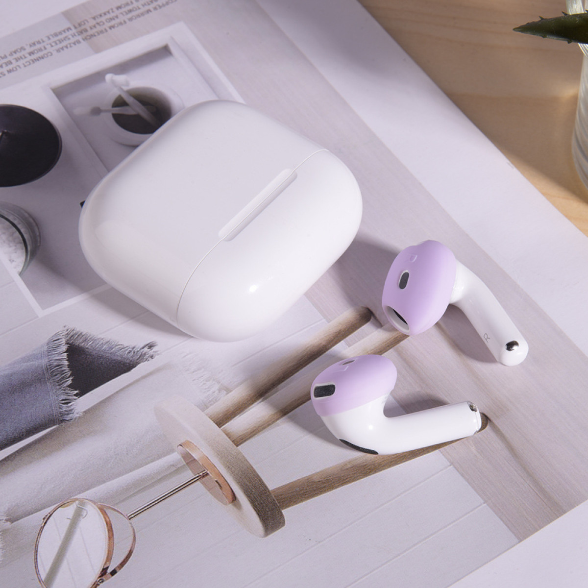 Weißes AirPods-Etui und zwei Ohrhörer mit lila Hüllen auf einer Zeitschrift. Weitere Objekte sind ein kleines Glas und ein runder, schwarzer Gegenstand.