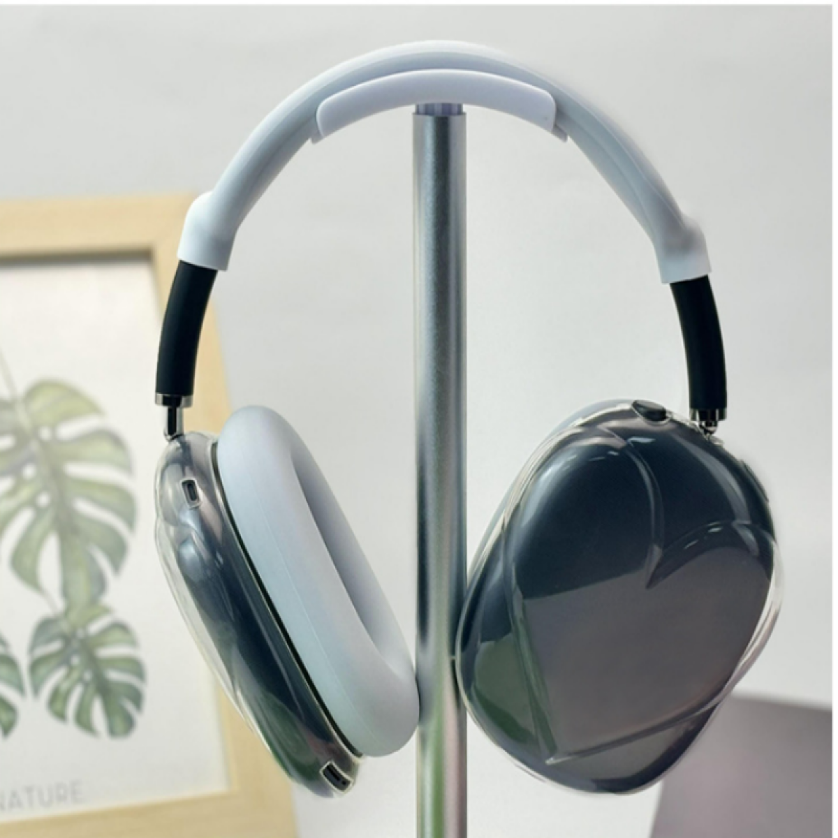 Słuchawki z białym pałąkiem i nausznikami, ze srebrnym stojakiem. Nauszniki są ciemnoszare. Ramka na zdjęcia jest w tle.