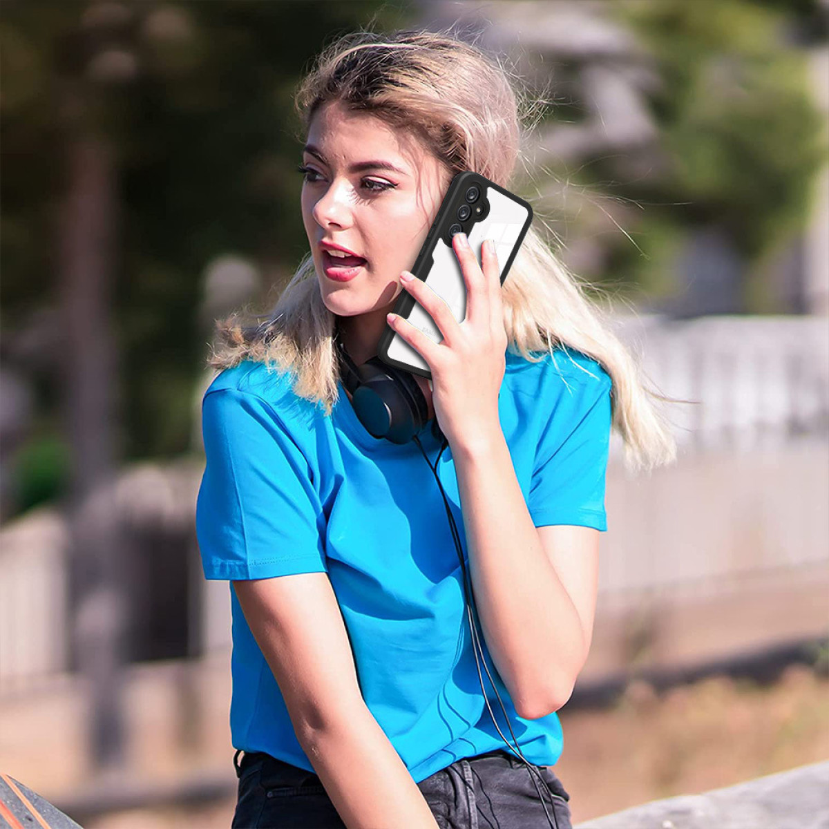 Una donna con i capelli biondi tiene un telefono all'orecchio. Indossa una camicia blu e le cuffie. Lo sfondo è verde sfocato.