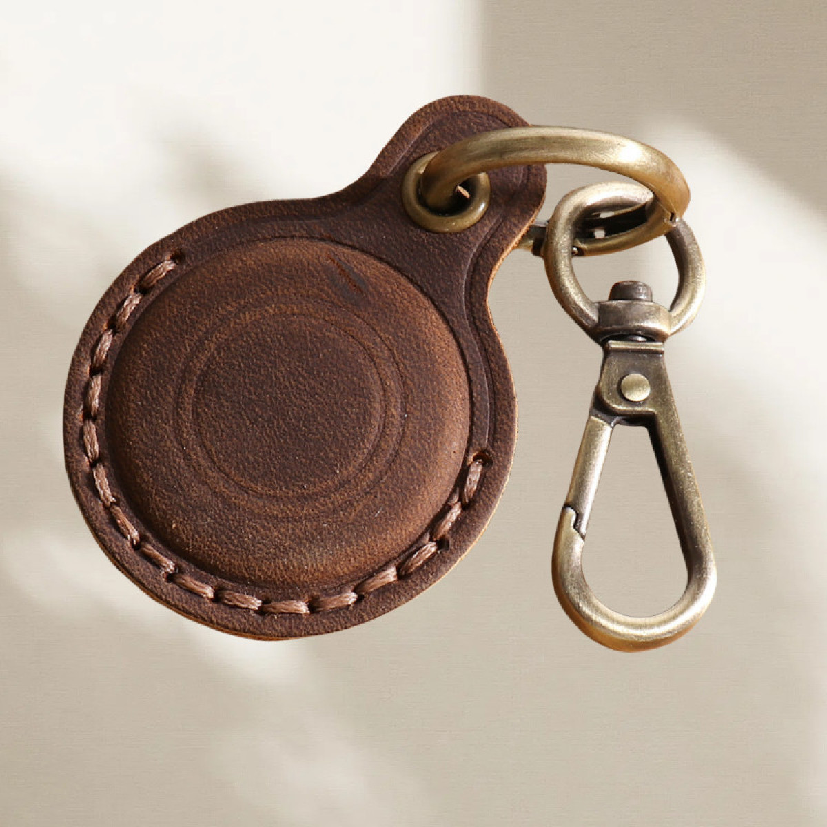 Ein brauner Leder-AirTag-Halter mit Metallverschluss und Ring vor neutralem Hintergrund. Der Halter hat genähte Kanten.