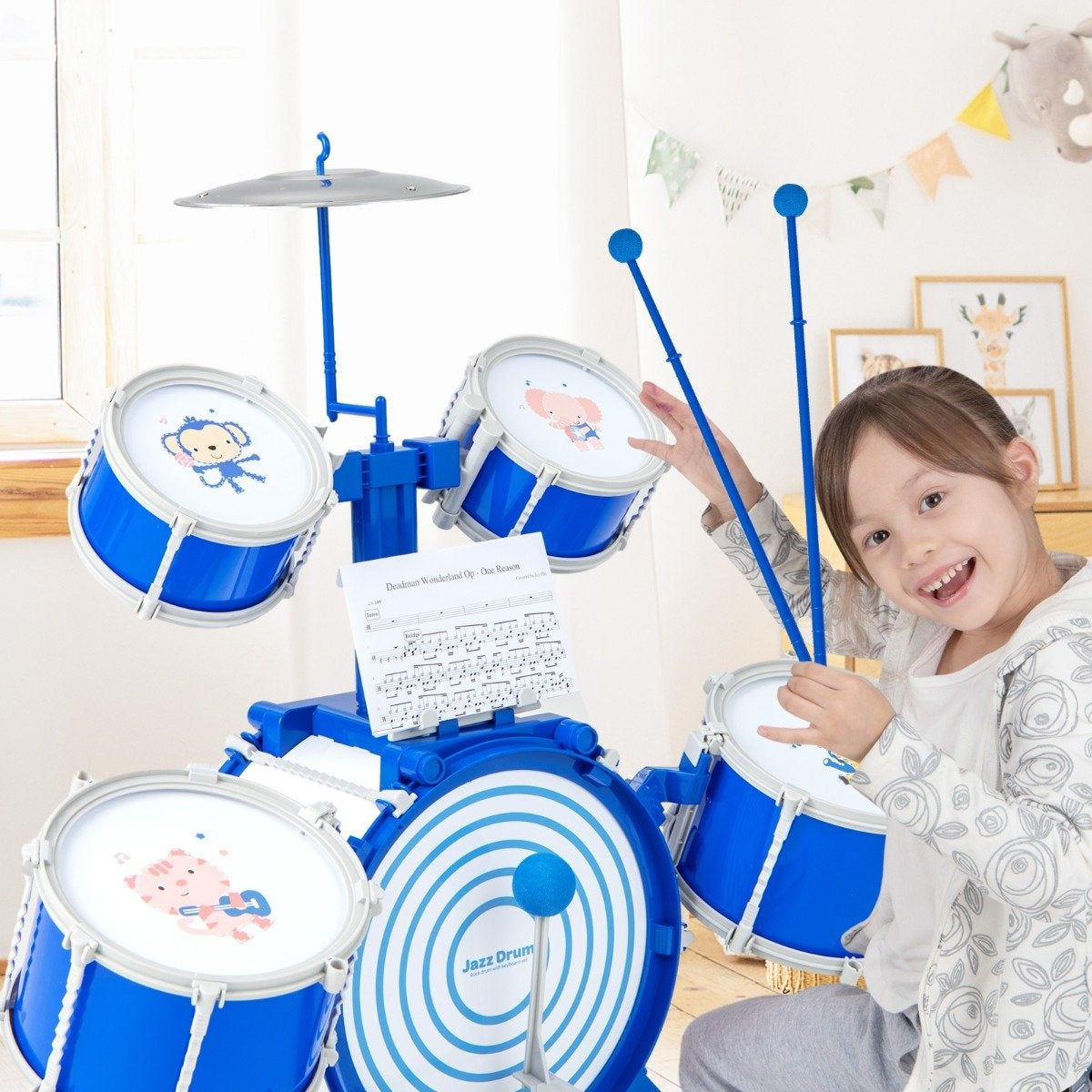 Una niña sonríe, tocando una batería de juguete azul con gráficos de animales. El set tiene un platillo, baquetas y partituras.