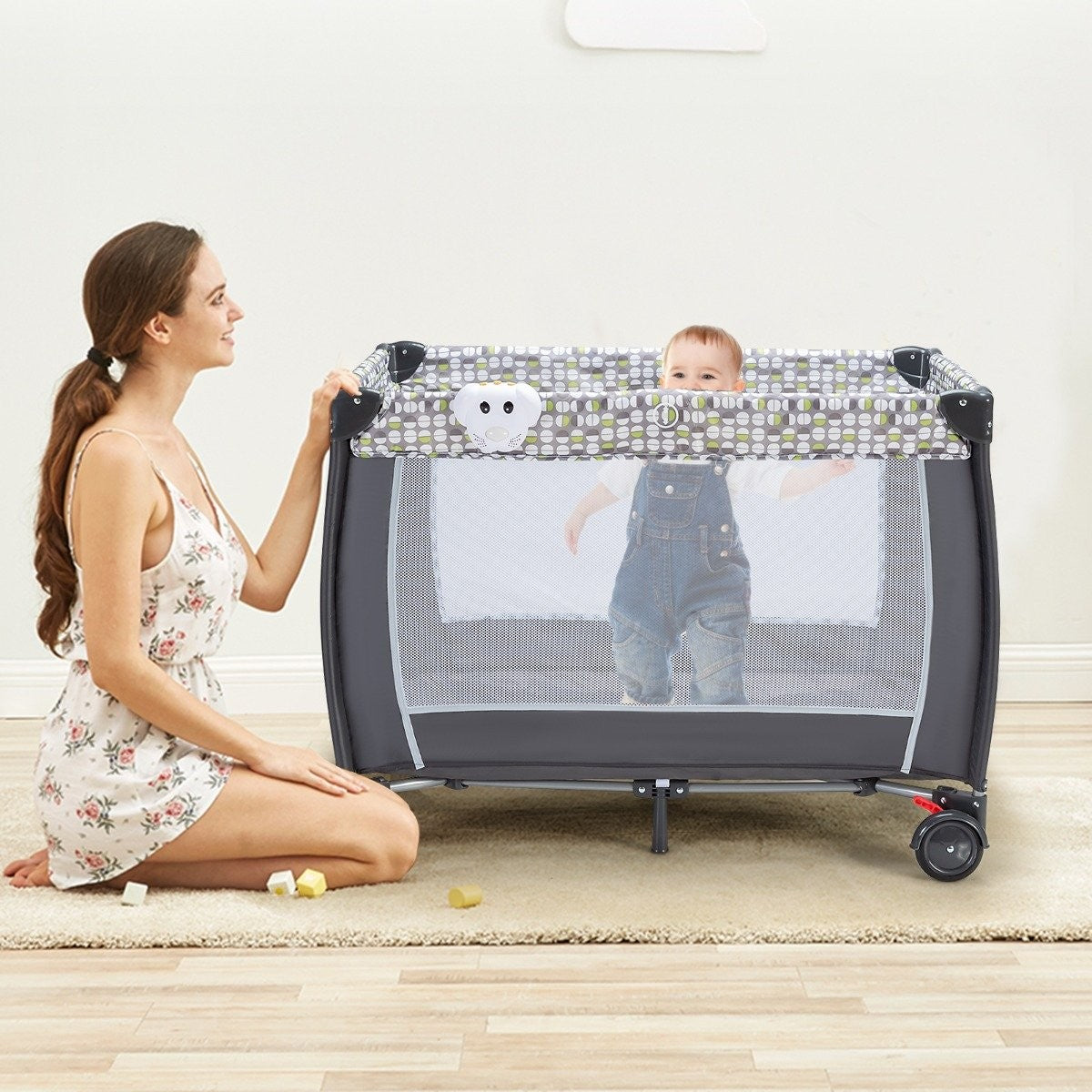 Een vrouw knielt bij een box met een baby erin. De baby draagt een blauwe overall. De box staat op een tapijt.