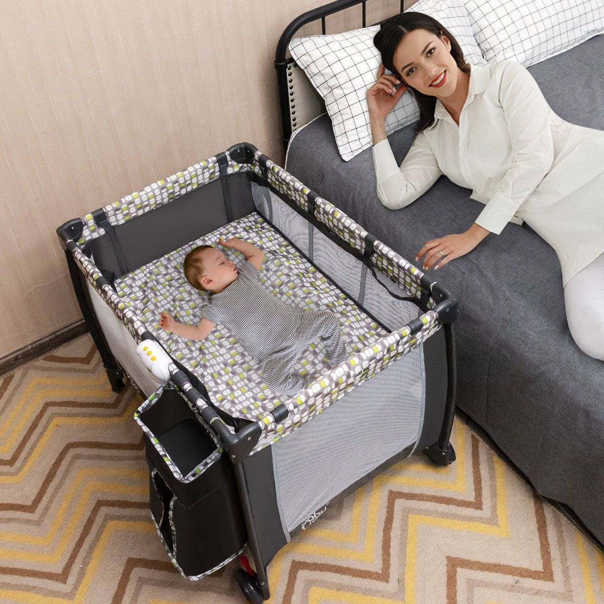 Een baby slaapt in een box. Een vrouw ligt in de buurt op een bed en glimlacht. De kamer heeft een patroonkleed.