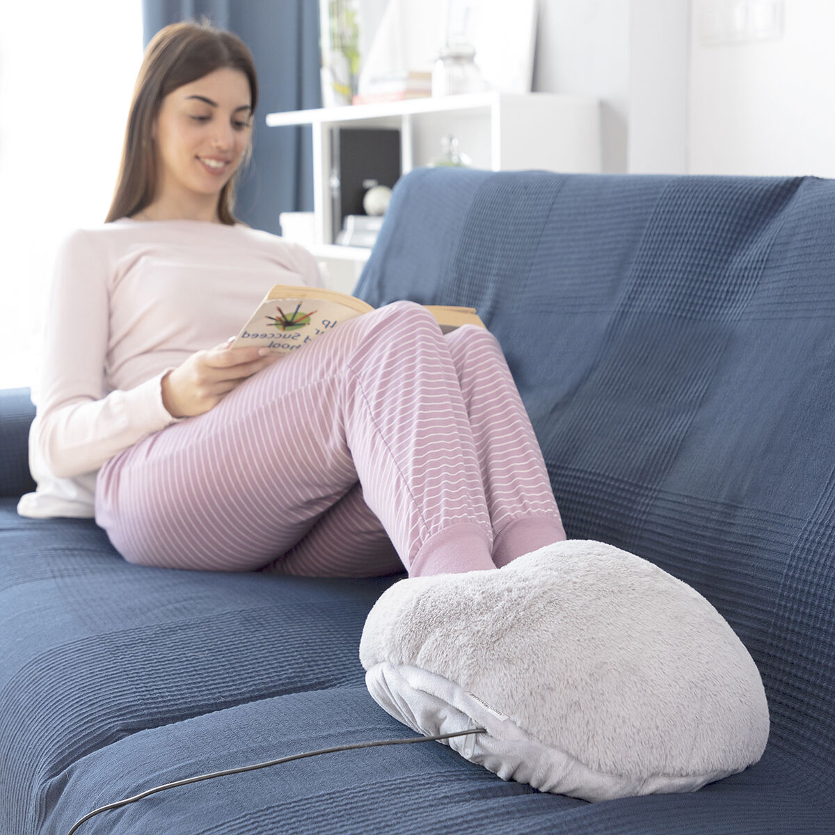 Mujer en un sofá azul lee con los pies en un calentador de pies. Lleva un pijama rosa. Fondo: estantería y ventana.