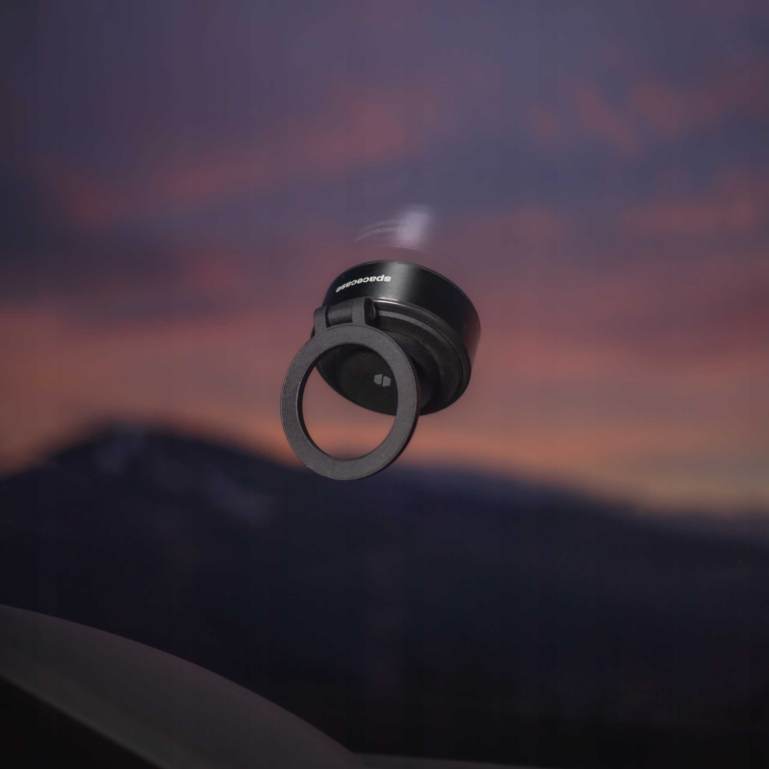 Czarny uchwyt na pierścień do telefonu z napisem 'spacecase' unosi się na tle zachodu słońca.