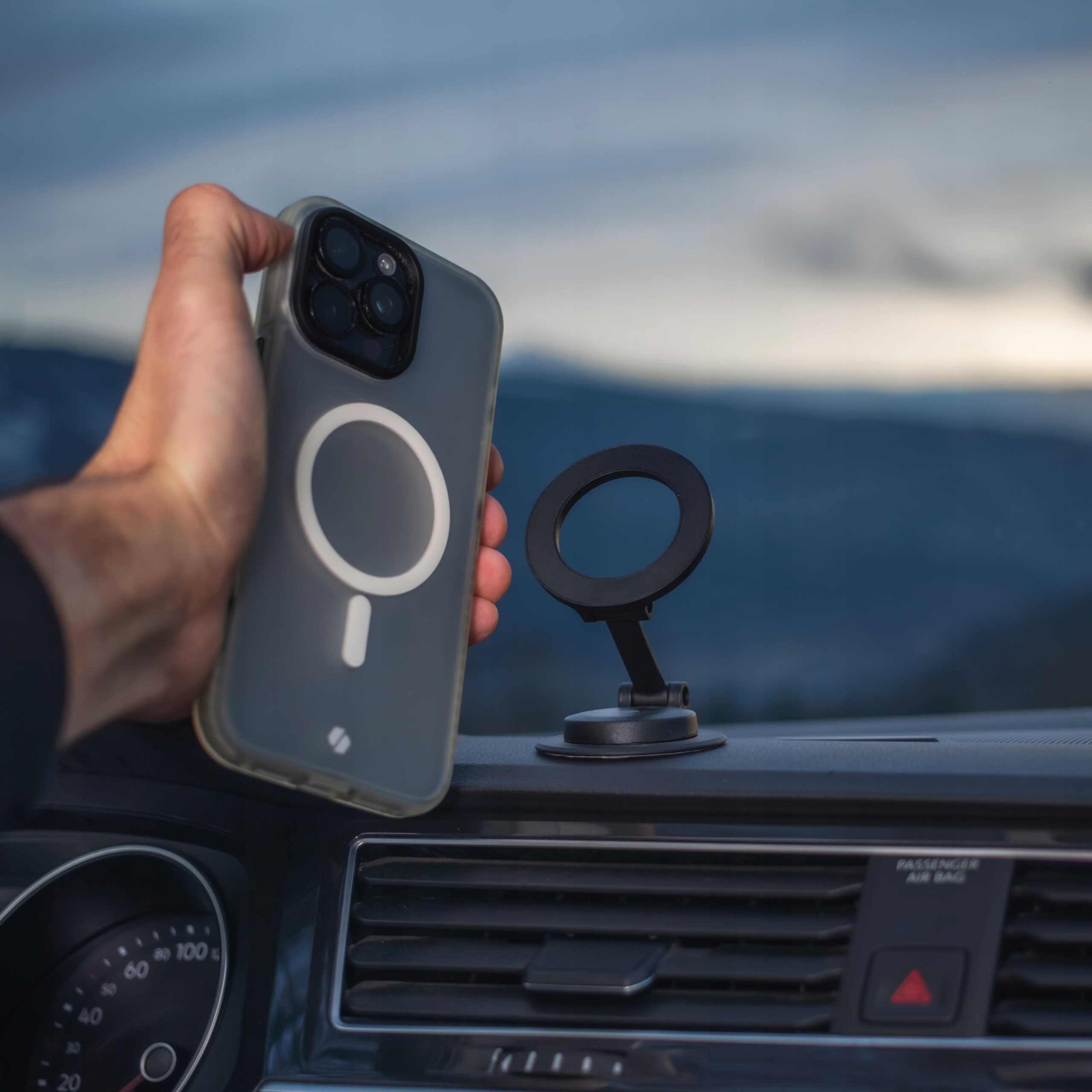 Dłoń trzymająca telefon w etui obok czarnego uchwytu samochodowego na telefon. W tle góry.
