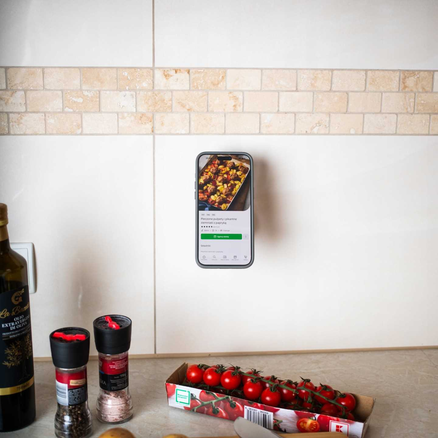 Telefon z przepisem na białej ścianie. Butelka oliwy z oliwek, młynki do pieprzu i pomidory na blacie.